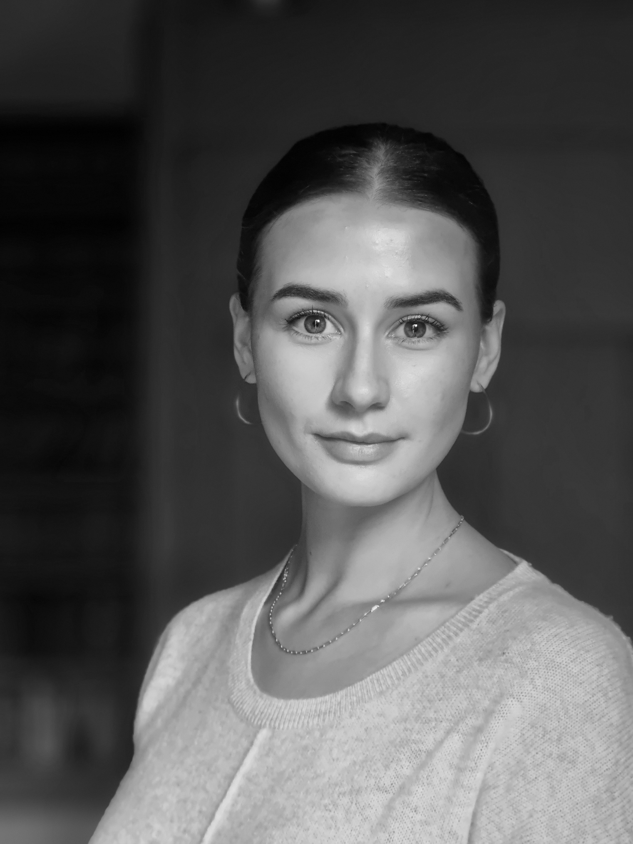 A black and white photo of a woman looking directly at the camera, with her hair pulled back, wearing hoop earrings, a necklace, and a light-colored top, against a dark background.