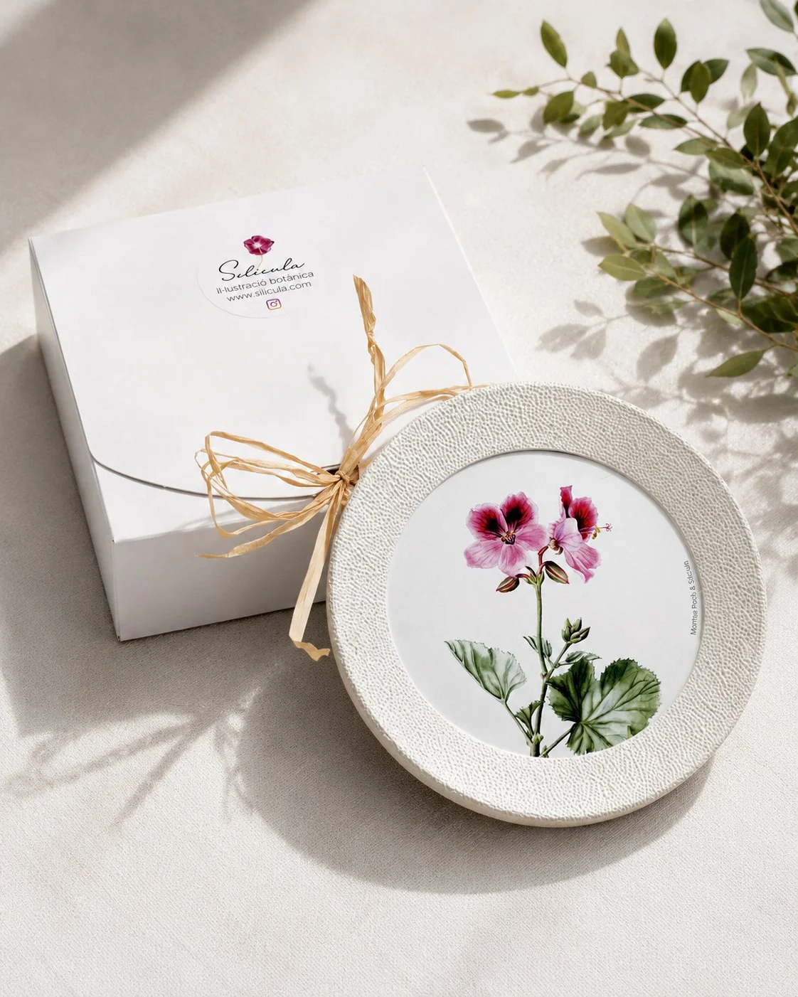 Caja blanca para packaging de un marco de la colección Miniaturas del jardín con ilustración en acuarela en su interior, decorados con una cinta de rafia y hojas verdes.