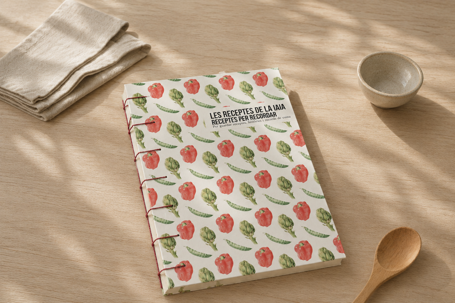 Libreta recetario con cubierta decorada con ilustraciones en acuarela de pimientos, alcachofas y judías, sobre una mesa de madera, junto a un bol, un cucharón de madera y un paño de cocina.