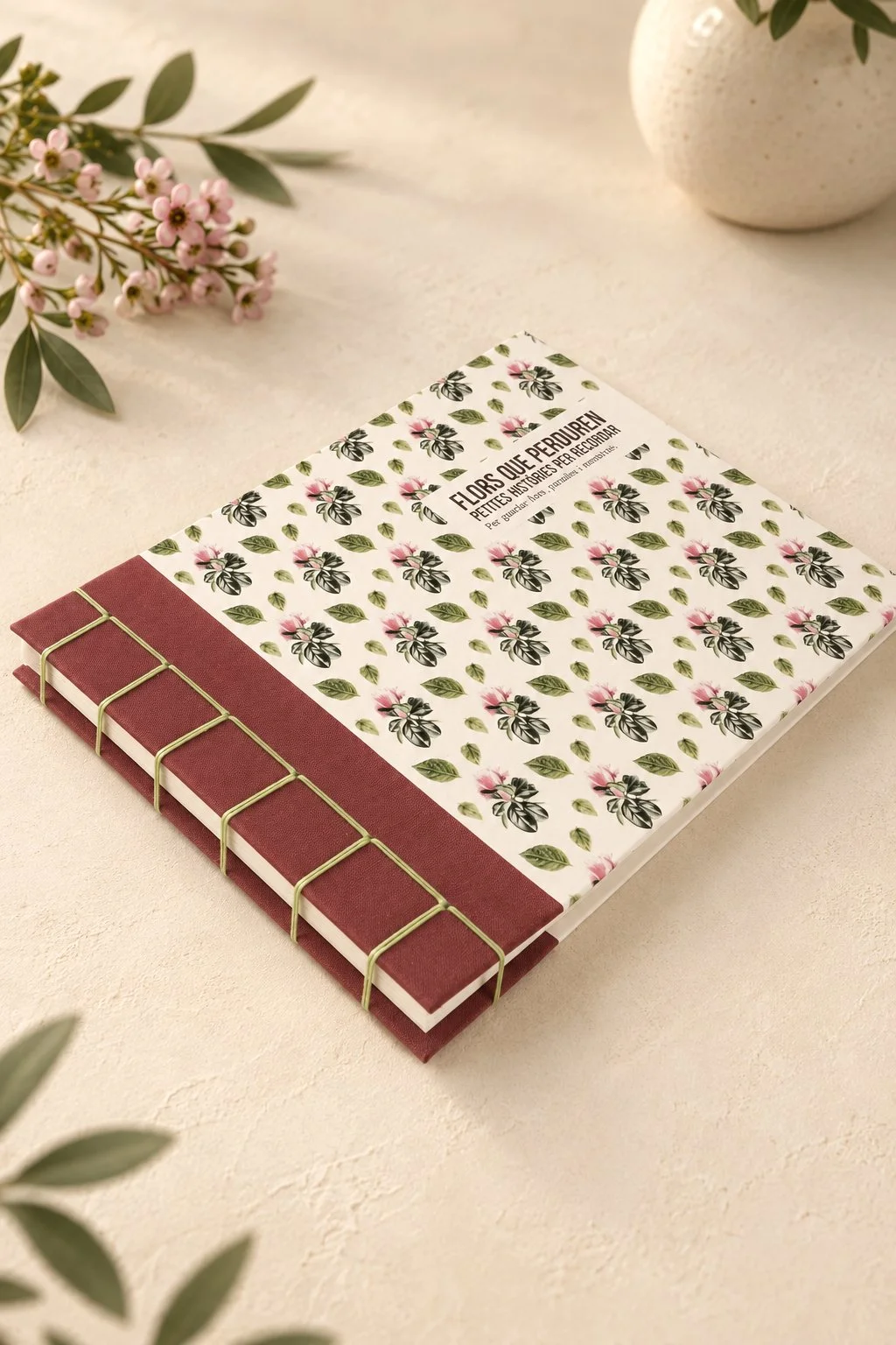 Libreta artesanal con cubierta decorada con motivos florales en acuarela, y con un lomo visto de tela granate, en una mesa con flores y una vasija blanca a su lado.