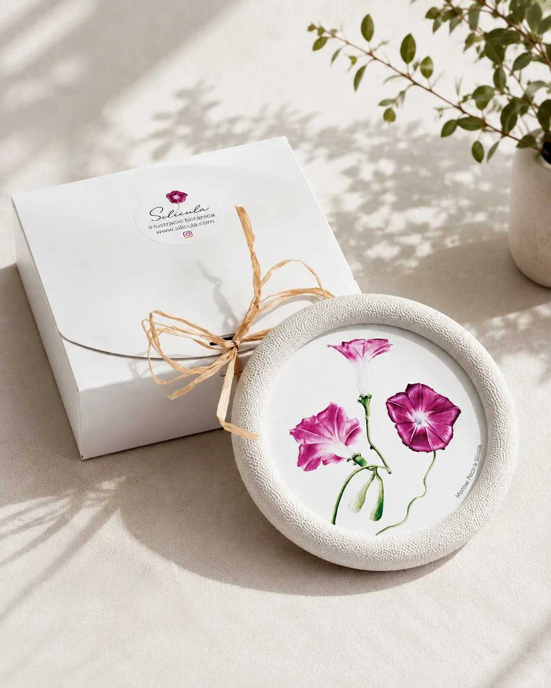 Caja blanca para packaging de un marco de la colección Miniaturas del jardín con ilustración en acuarela en su interior, decorados con una cinta de rafia y hojas verdes.