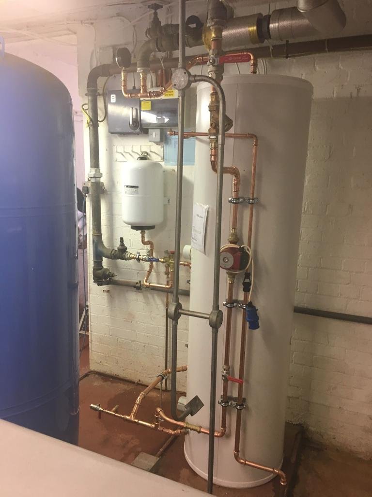Industrial hot water storage tank with copper piping and valves in a utility room.