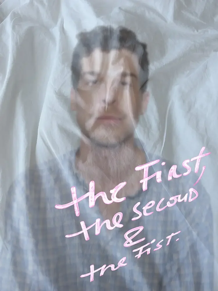 A man with dark hair and facial hair is behind a semi-transparent plastic sheet with pink handwritten text that says 'the First, the Second & the First'.