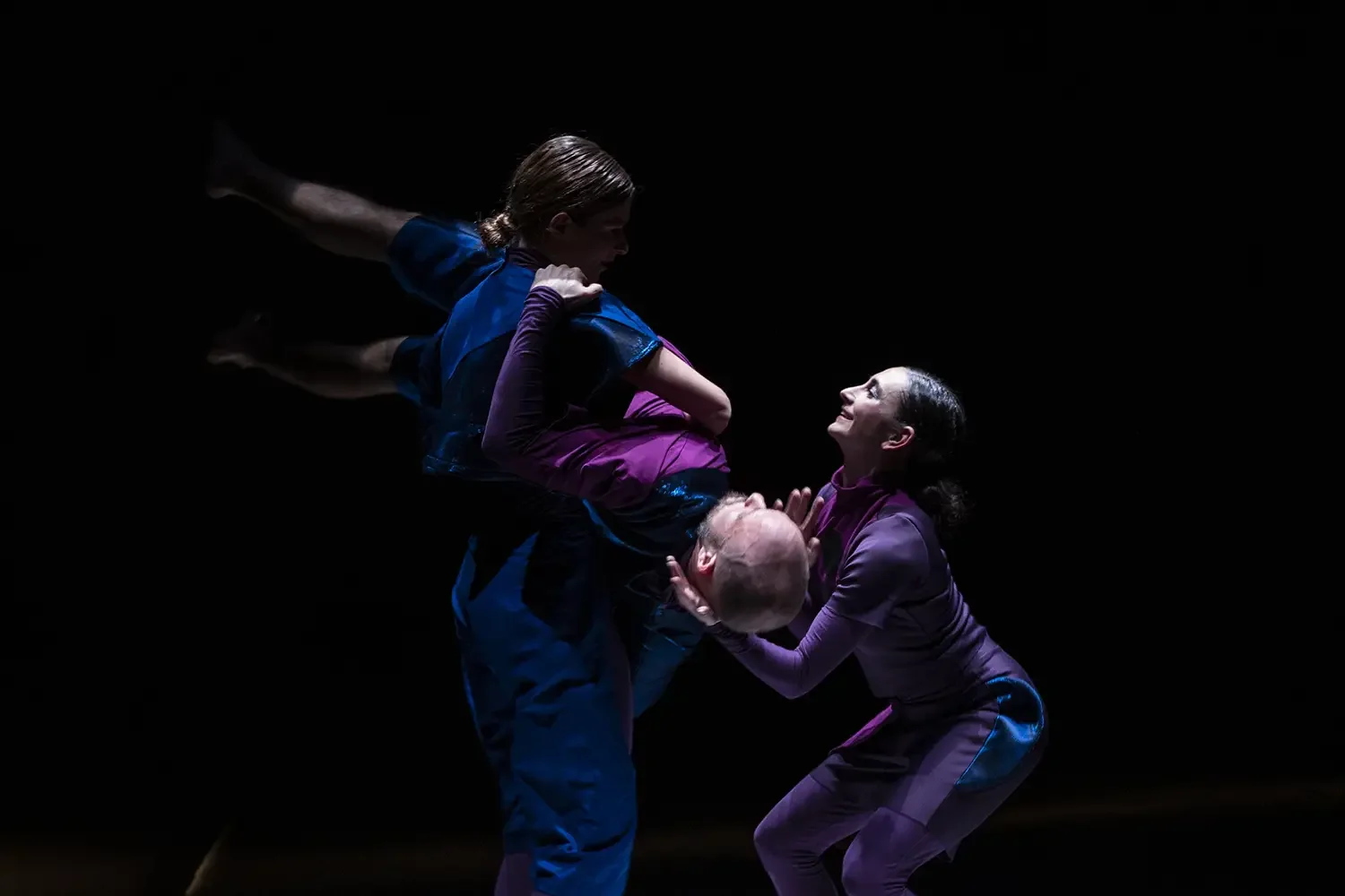 Three performers with colorful outfits performing a dance or acrobatic routine on a dark stage, with dramatic lighting