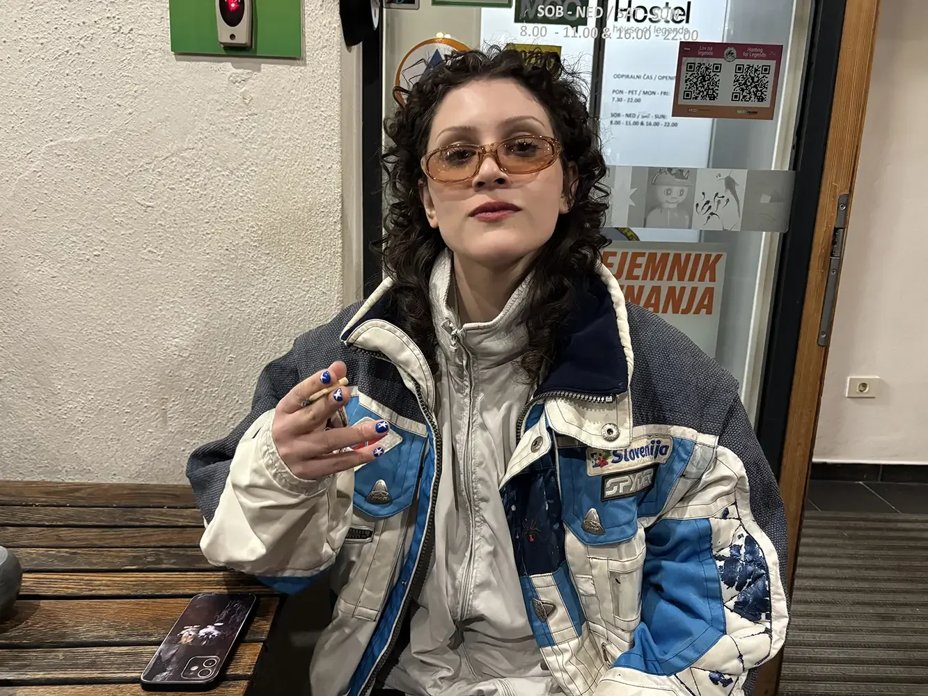 A woman with curly hair wearing sunglasses, a white and blue jacket, and holding a blue and white nail polish, sitting at a wooden table with a phone on it, in front of a textured wall and a glass door with posters and signs.