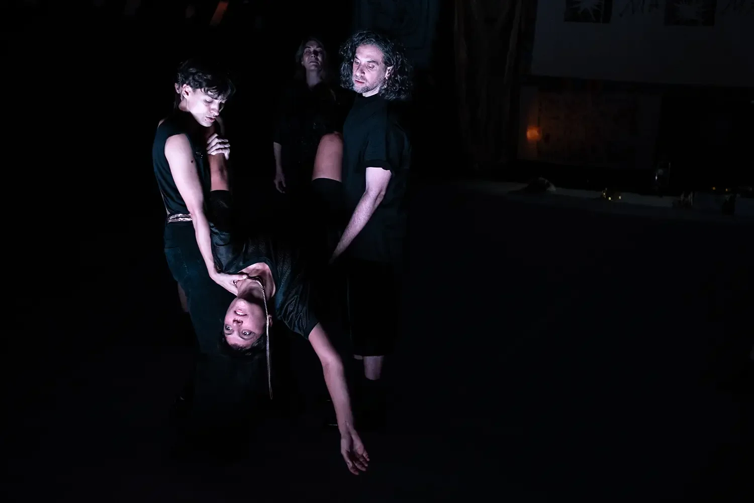 Group of five people in a dark setting, one person is upside down with their head hanging towards the ground, others stand around watching.