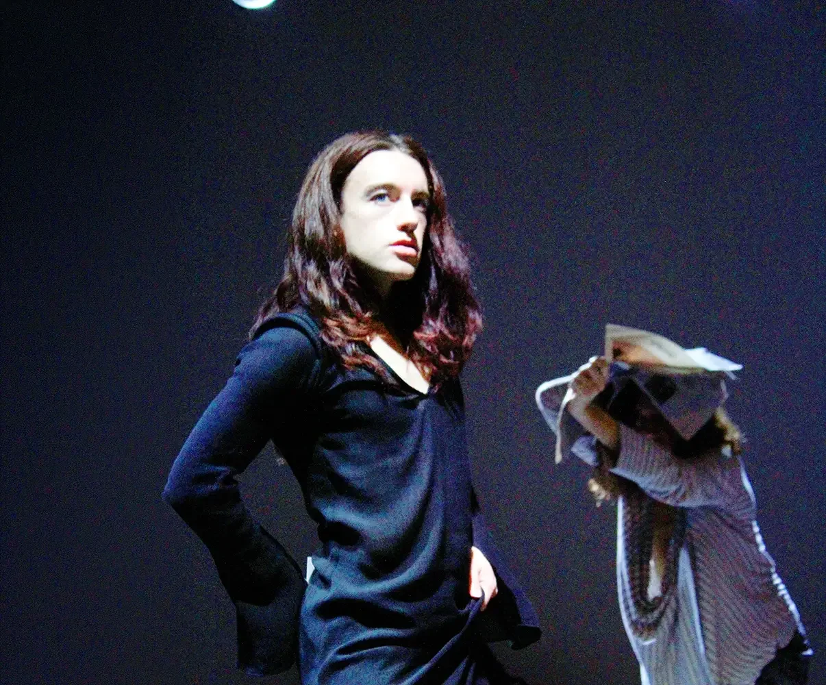 A woman with long brown hair wearing a black top, standing against a dark background, looking off to the side.