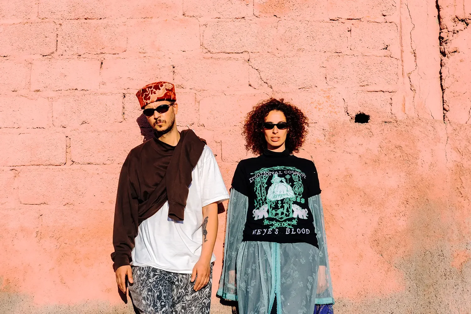 Two young adults standing in front of a pink brick wall indoors or outdoors, wearing fashionable clothing and sunglasses.