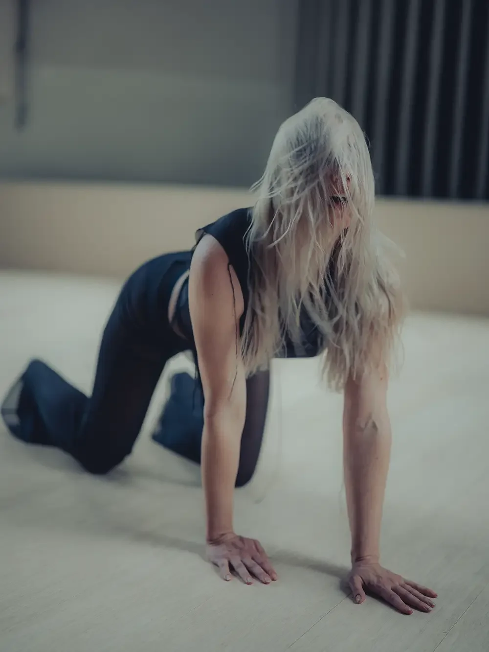 A woman with long, wet, blonde hair, wearing a black sleeveless top and black pants, on her hands and knees on the floor, with her hair obscuring her face.