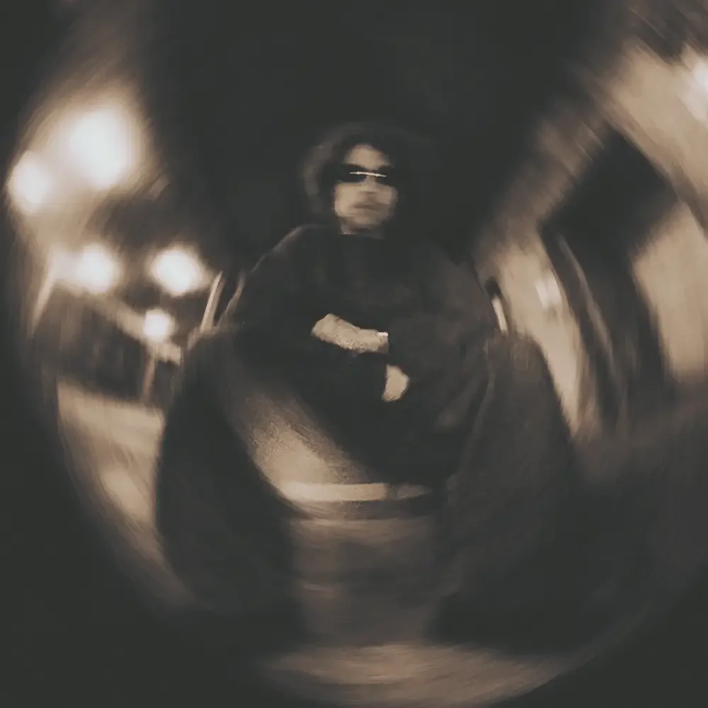 Person wearing sunglasses and dark clothing, sitting in a reflective, distorted environment, possibly a metallic surface or a mirror.