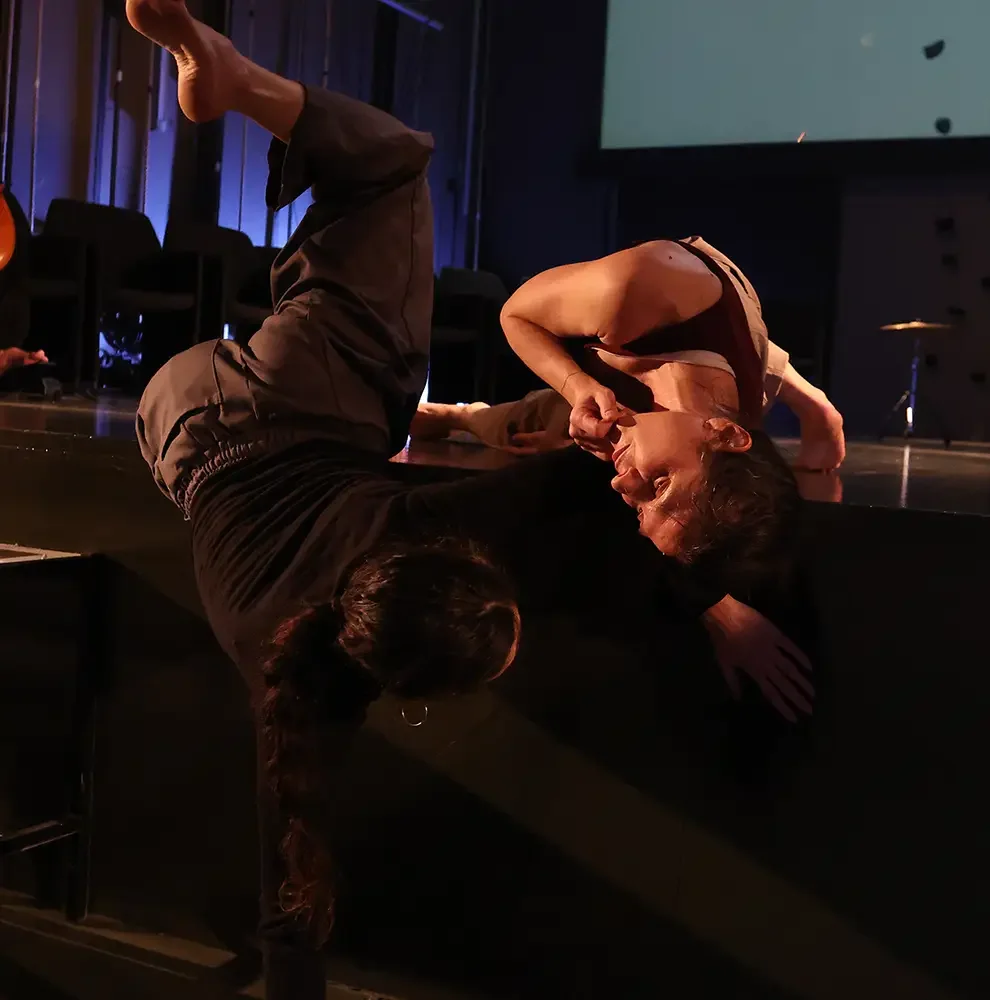 Two women are practicing acro yoga or a similar partner yoga move on a dark stage. One woman is lying on her back with her legs raised, while the other woman is leaning over her, supporting her head with one hand and holding her ankle with the other hand.