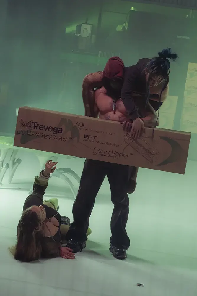 A woman on the floor reaching up with one hand, while a man stands holding a large cardboard box labeled with various brand names, in an indoor space with a greenish hue and gym equipment in the background.