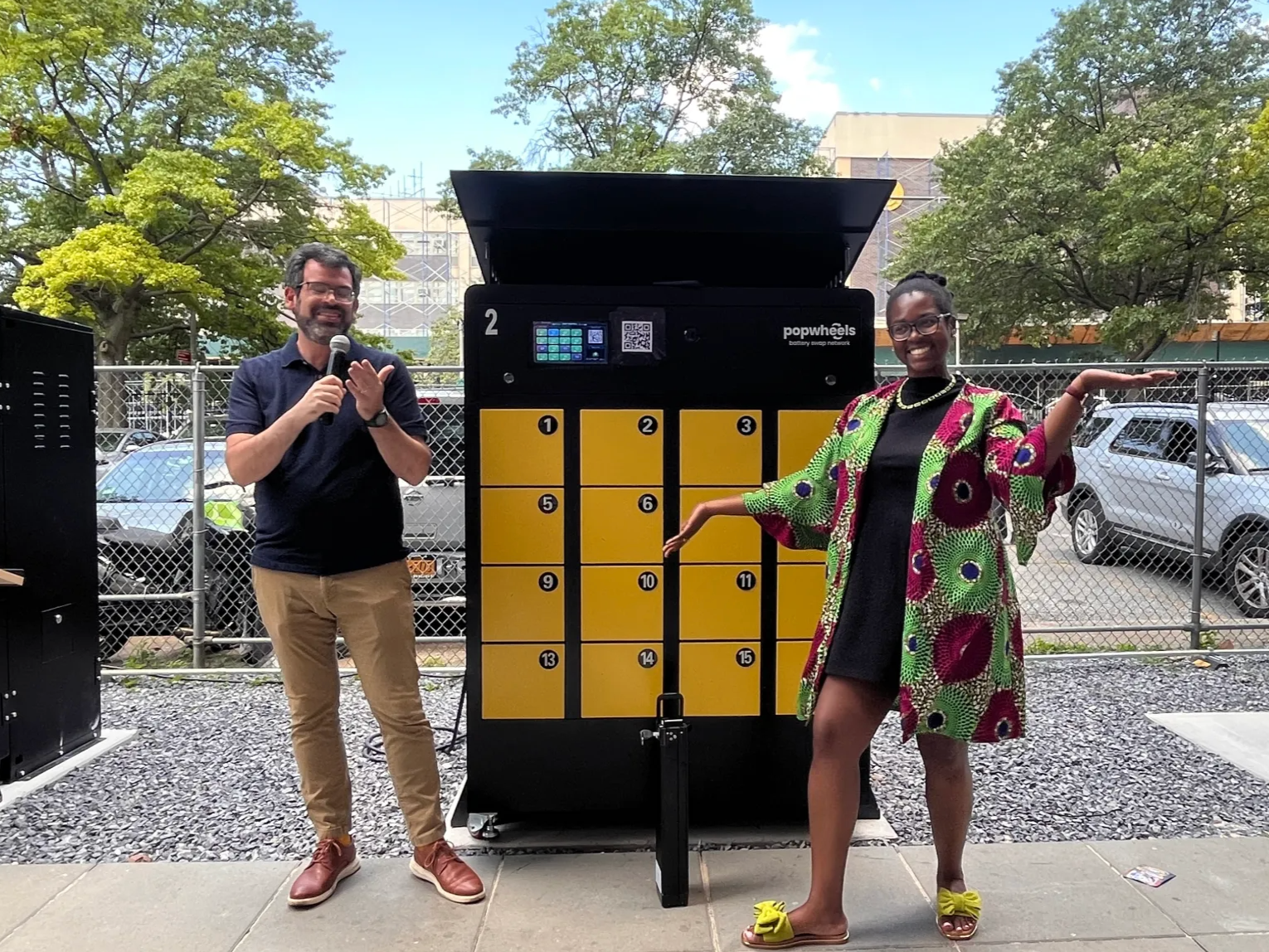 Two people standing outdoors, posing next to a large yellow and black battery swapping station with a screen. One person is a man holding a microphone, and the other is a woman with arms extended, smiling.