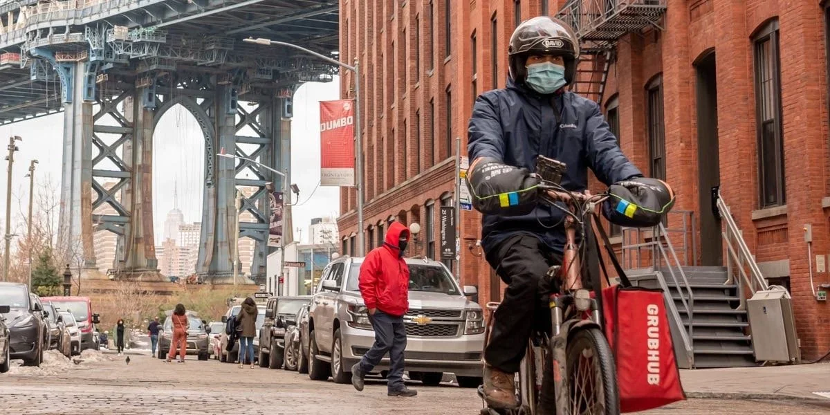 A person riding a bicycle on a city street wearing a mask, helmet, and dark jacket, with a large red bag labeled 'GRUBHUB' attached to the bike. Several pedestrians and parked cars line the street, with a large bridge and NYC skyline in the background.