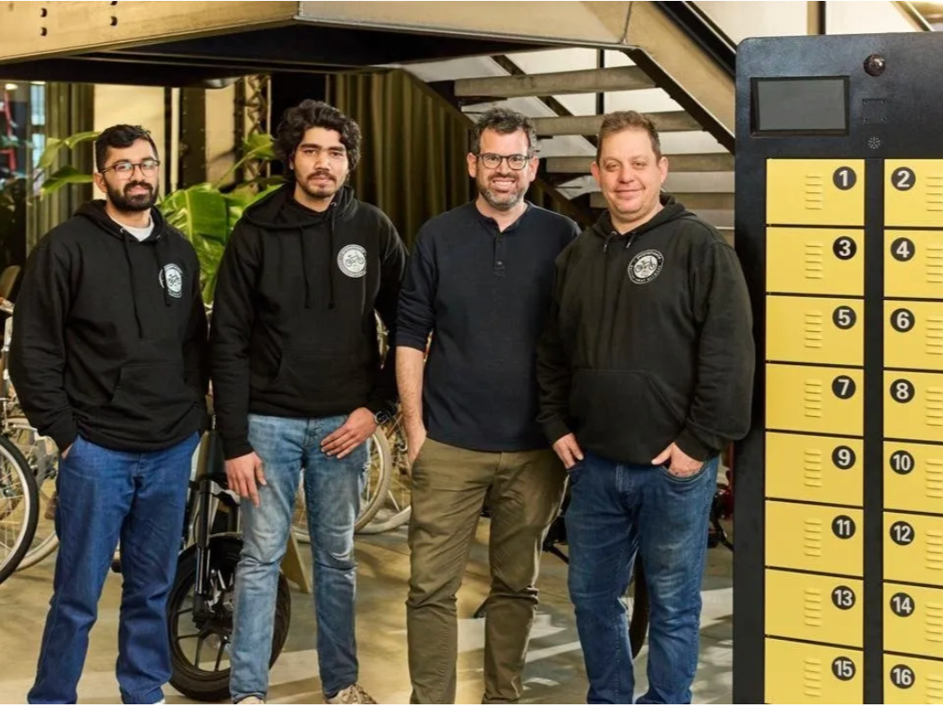 Four men standing indoors, some wearing black hoodies with a logo, near a yellow lockers and bicycles.