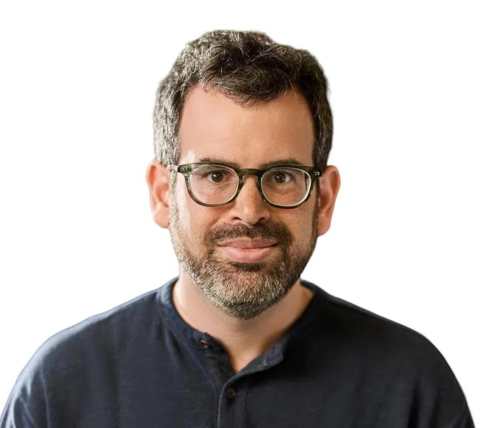 A man with short dark hair, glasses, and a beard, wearing a dark collared shirt, posing against a plain white background.