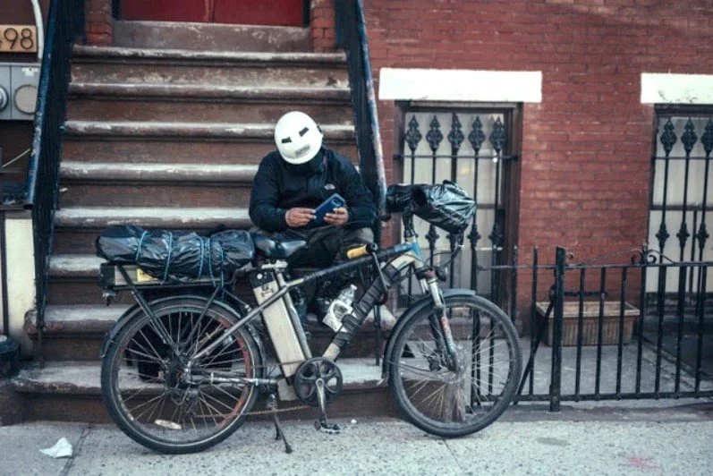 Person wearing a white helmet sitting on stairs with a bicycle parked beside them, looking at a device, in front of brick buildings with metal railing.