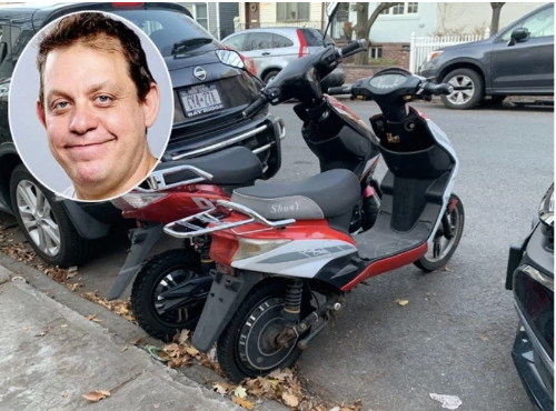 A red and black electric scooter parked on a street with other cars in the background. An inset image of a smiling man's face is in the top left corner of the photo.
