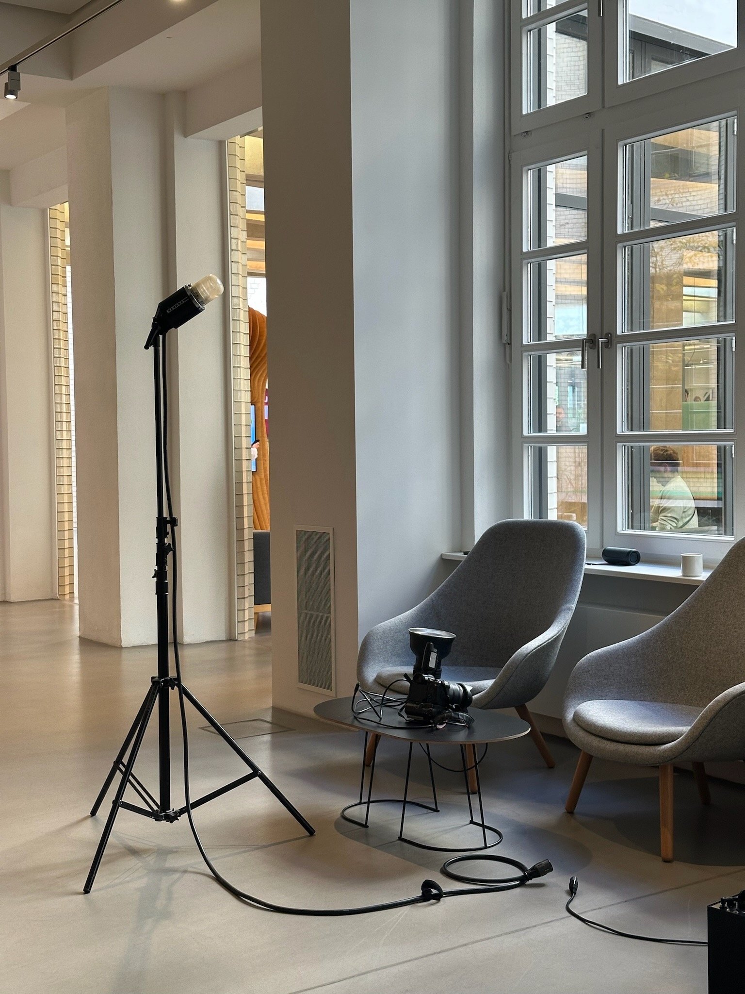 Videodreh, behind-the-scenes, contentproduktion, Aufnahmebereich mit zwei grauen Sessel vor einem großen Fenster, Kamera und Beleuchtung auf einem Tisch, Kabel auf dem Boden, modernes Interieur.