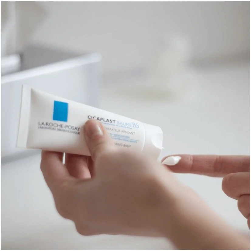 Eine Hand hält eine weiße Tube der La Roche-Posay Cicaplast Baume B5 vor einem weißen Hintergrund, während die andere Hand auf den Finger zeigt.