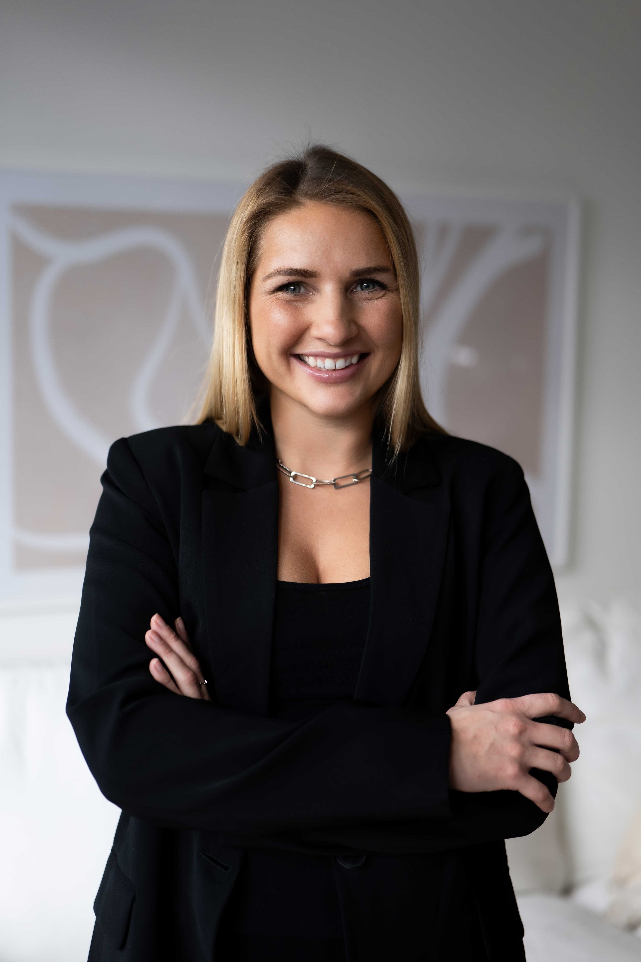 Eine lächelnde Frau mit blonden Haaren, die eine schwarze Jacke trägt, in einem modernen Raum.