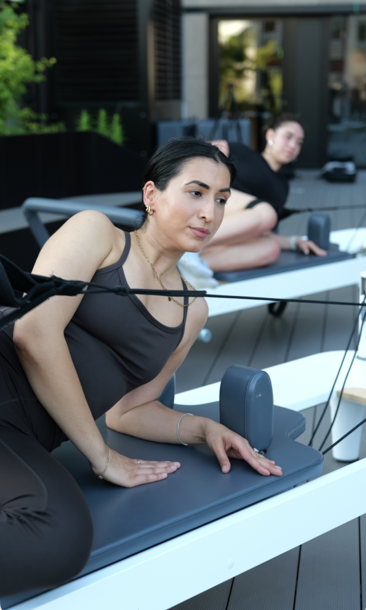 Zwei Frauen, eine vorne, eine im Hintergrund, beim Pilates auf einer Terrasse mit Blick nach draußen.