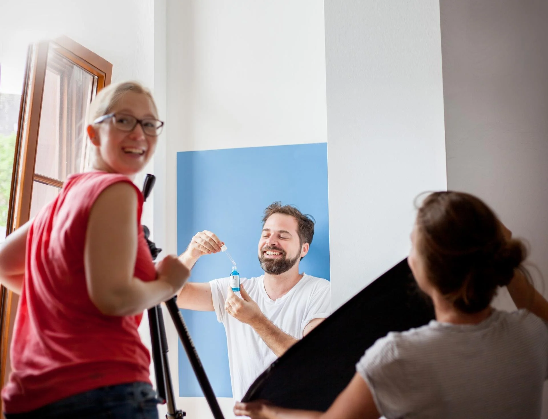 Drei Personen beim Fotoshooting, eine Frau hält einen Regenschirm, eine sitzt im Rollstuhl und bekommt eine Flasche mit Tropfen, während ein Mann lächelnd im Hintergrund steht.