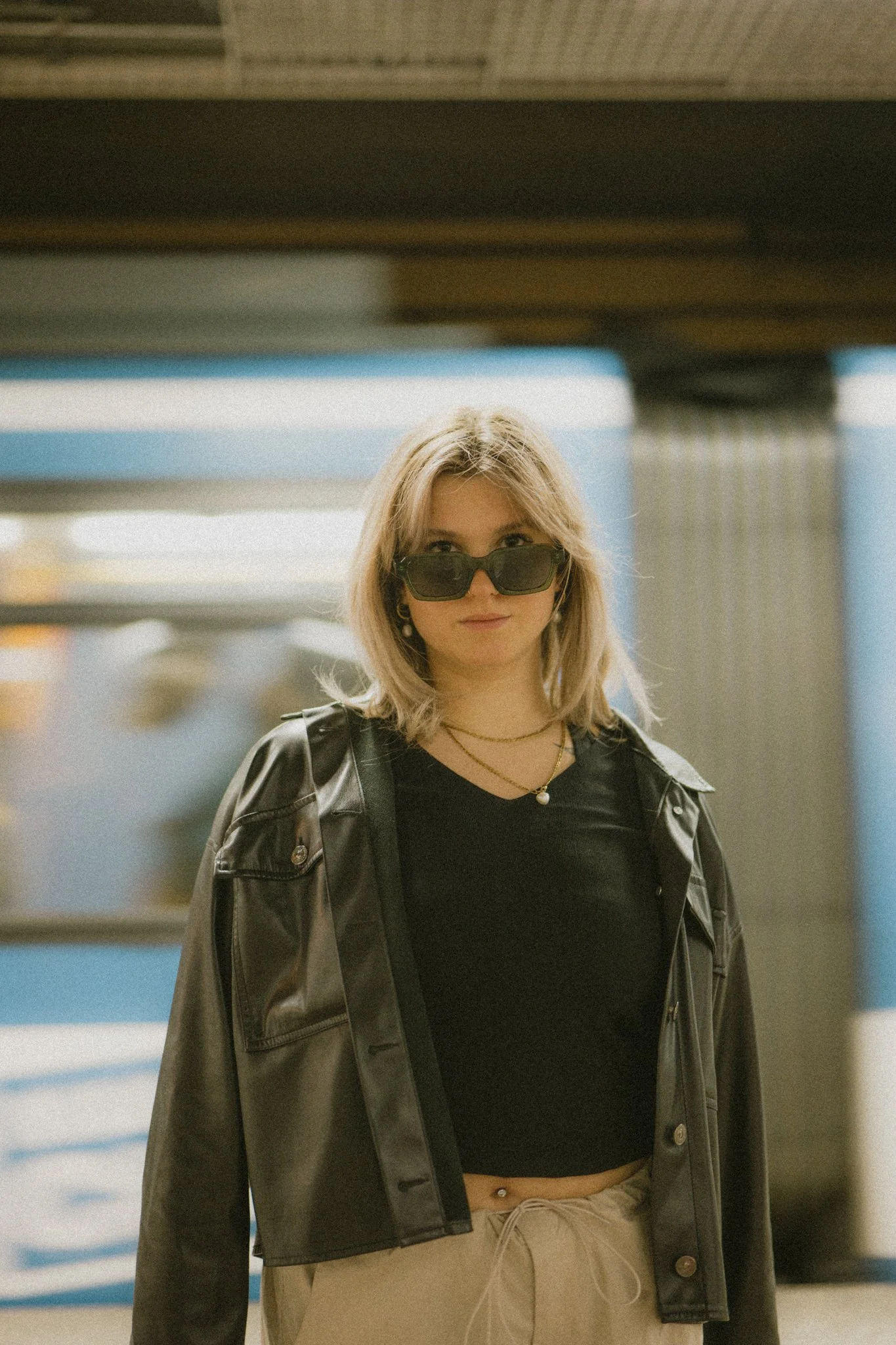 Fashion Shooting, Junge Frau mit dunkler Sonnenbrille, schwarzem T-Shirt, Lederjacke und beigen Hosen steht in einer U-Bahn-Station.