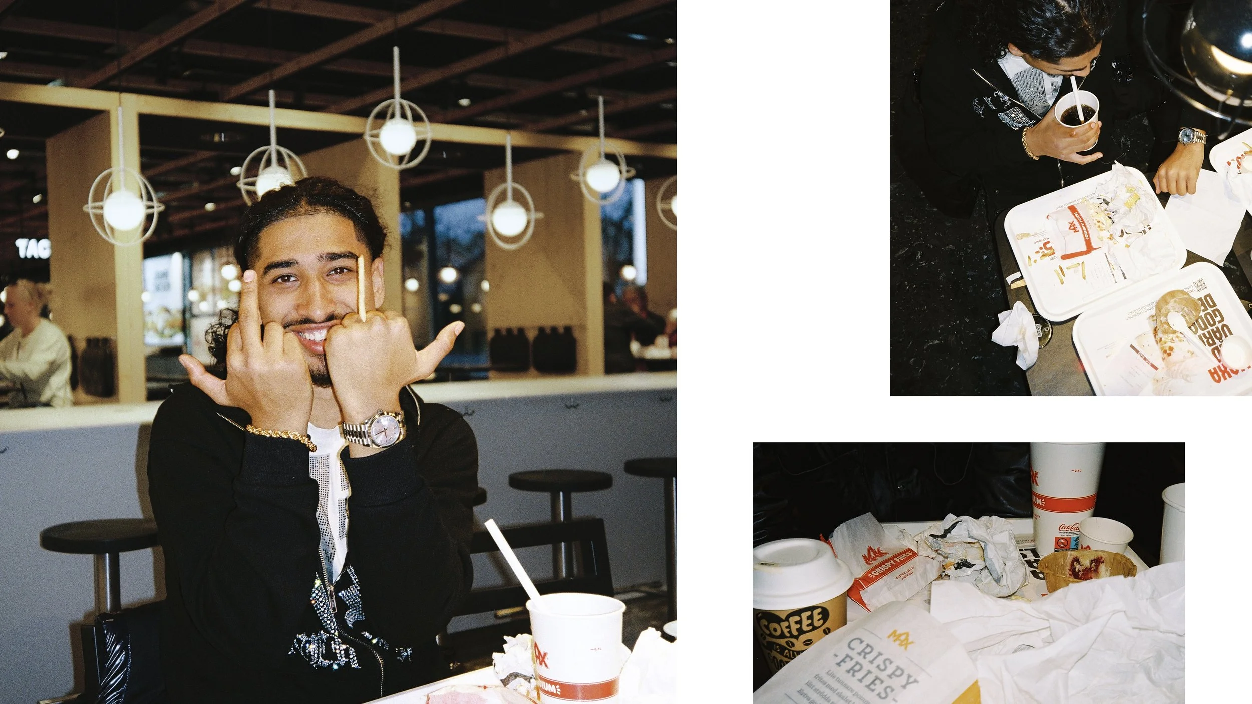 A man sitting at a table in a fast food restaurant, making a hand gesture with both hands, smiling, with food wrappers and drinks on the table. The restaurant has a modern interior with hanging lights.