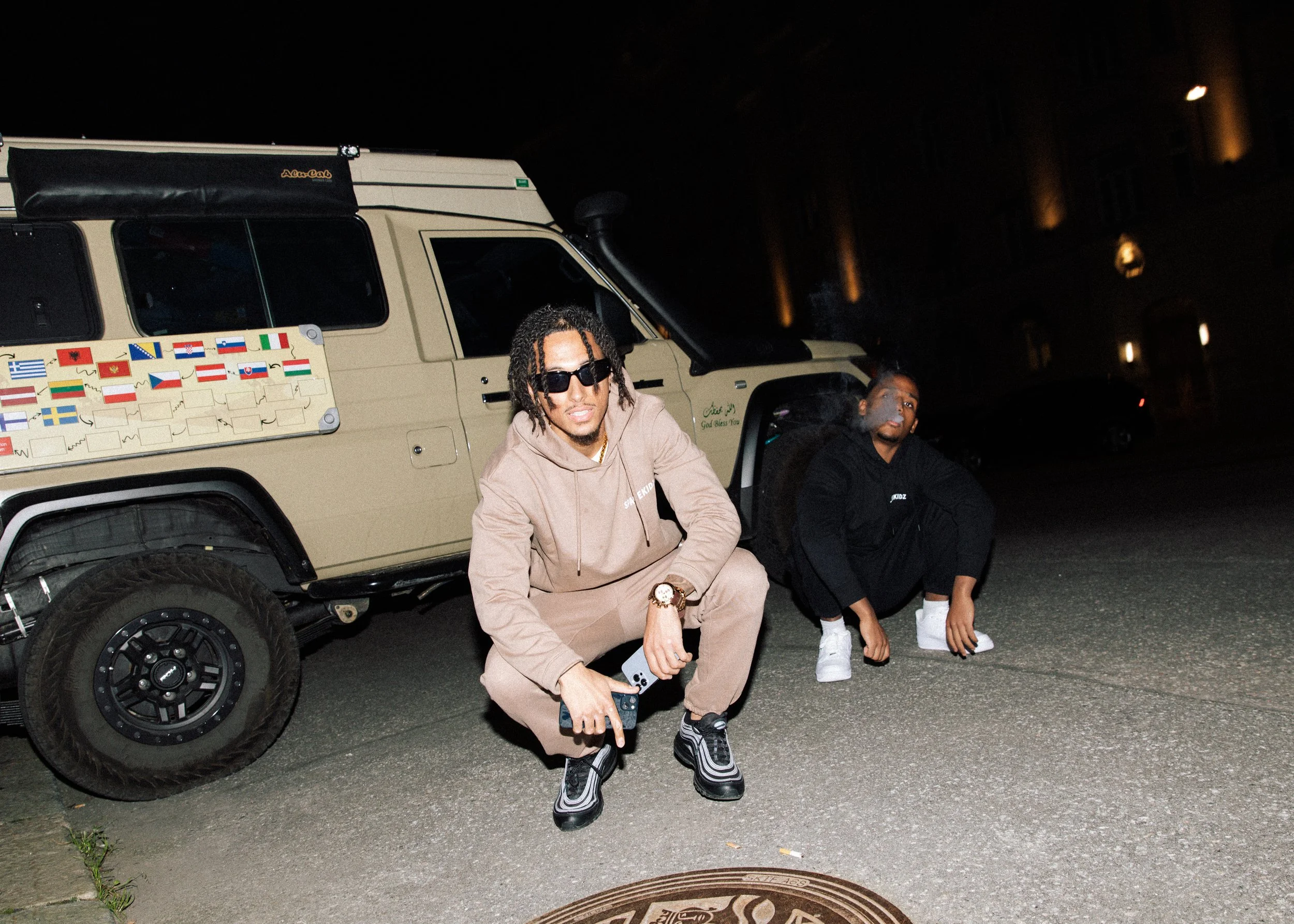 Two young men crouching on the street at night, one wearing a beige hoodie and sunglasses, the other in a black hoodie, in front of a beige off-road vehicle with flags on the side, under streetlights.