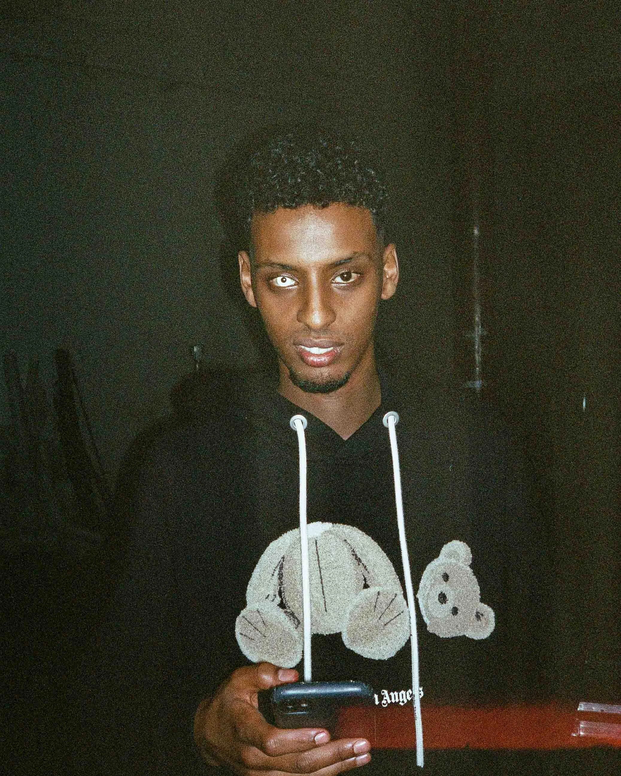 Young man with curly hair, wearing a black hoodie with a teddy bear graphic, holding a smartphone, in a dark room.
