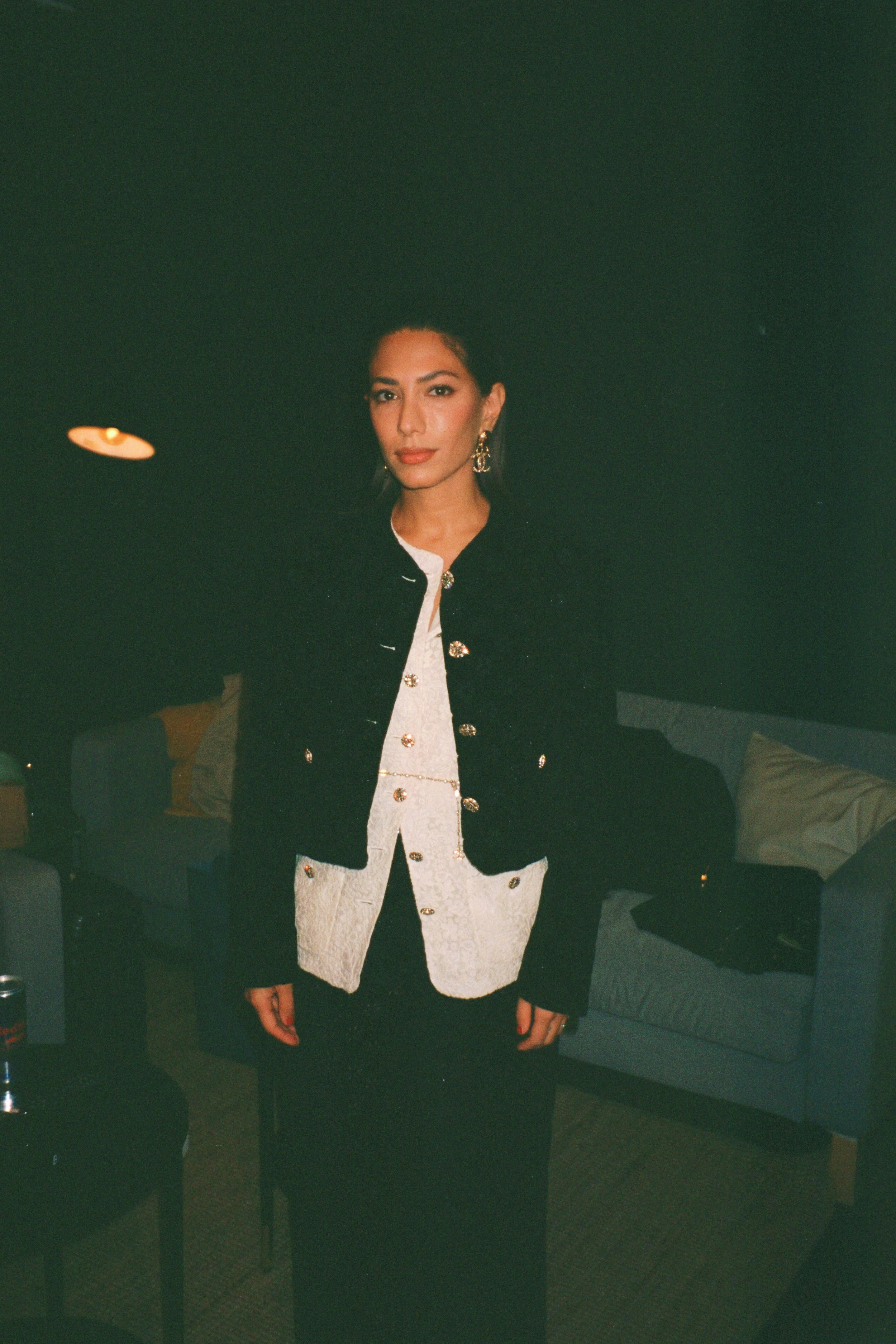 Woman standing in a dimly lit room, wearing a black jacket with gold buttons over a white top with gold embellishments, gold earrings, and dark pants.
