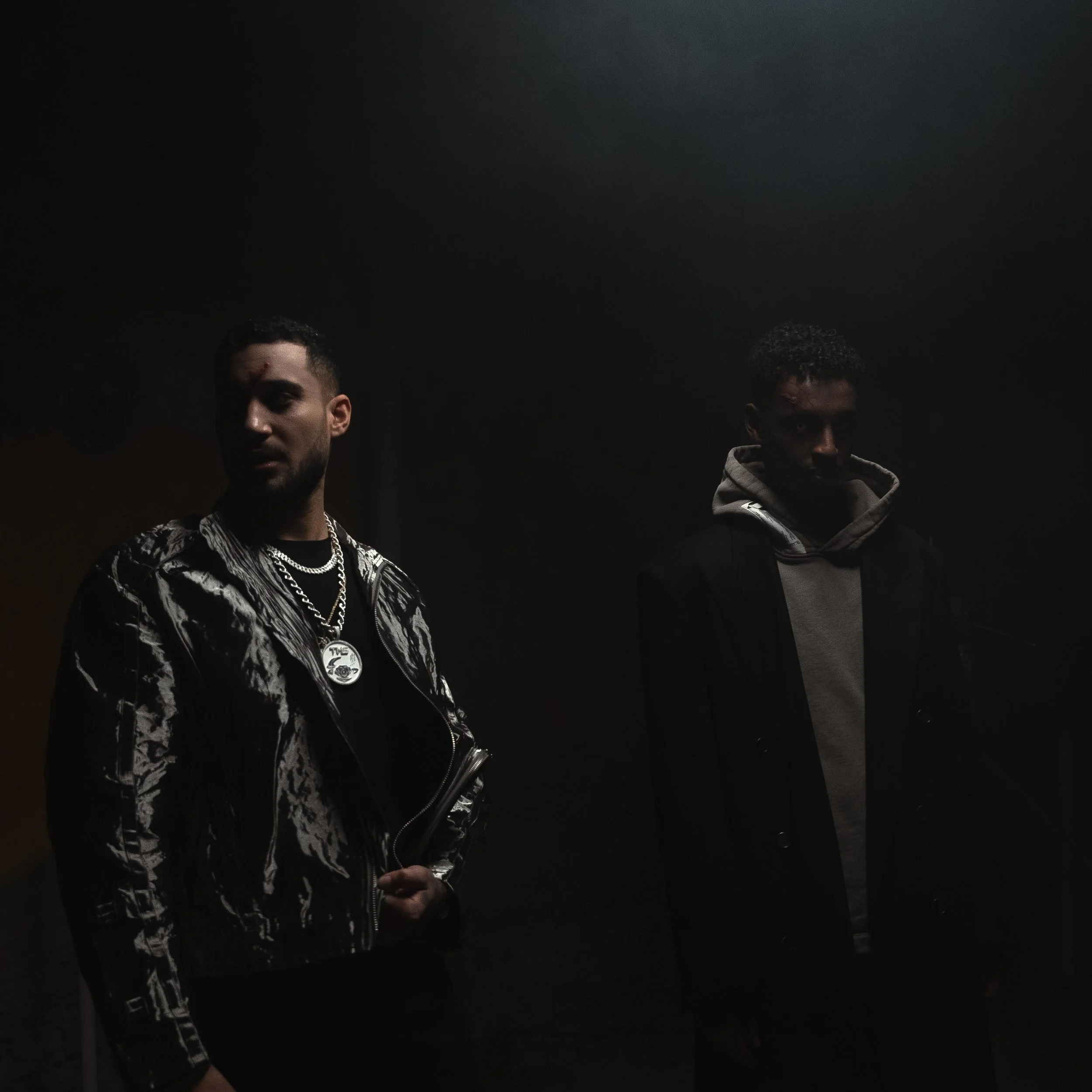 Two young men standing in a dark room, illuminated from above, with serious expressions. One has a beard, jewelry, and a patterned jacket; the other wears a hooded sweatshirt and coat.