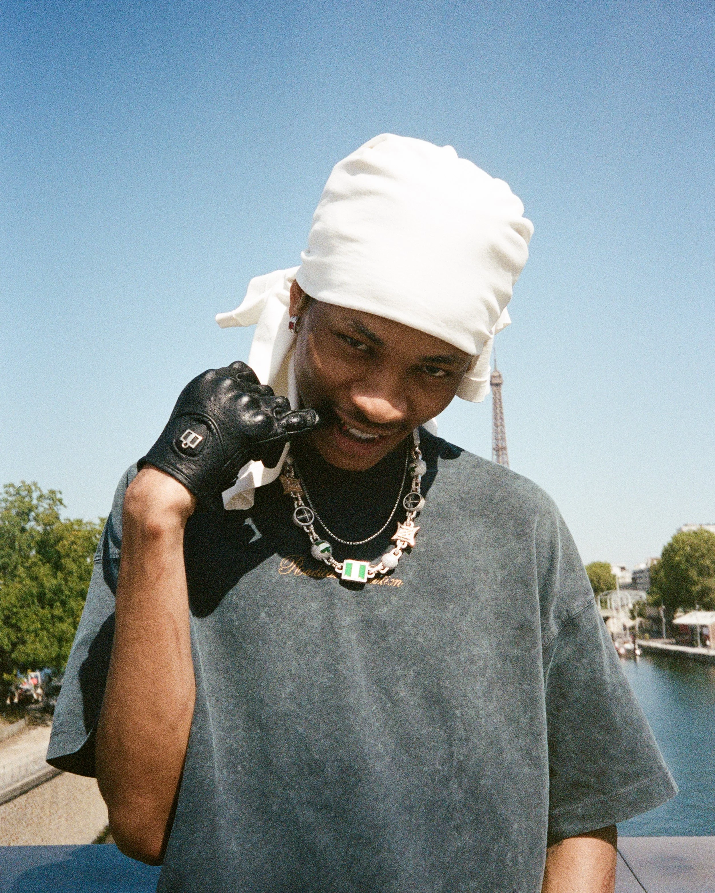 A person wearing a white headwrap, black glove, and layered necklaces, standing outdoors near a river with trees and buildings in the background.