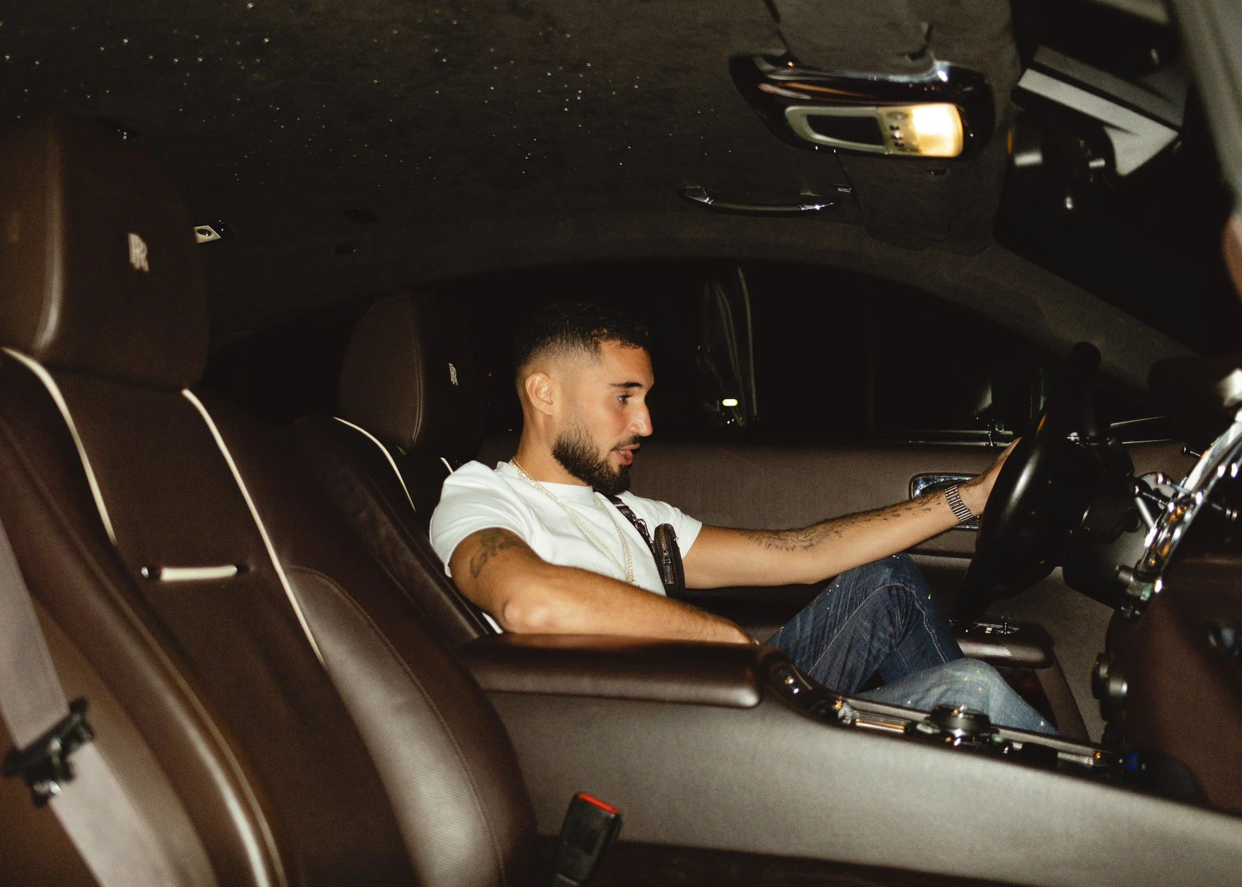 A man with tattoos and a watch sitting in the driver's seat of a luxury car at night, looking at the dashboard.