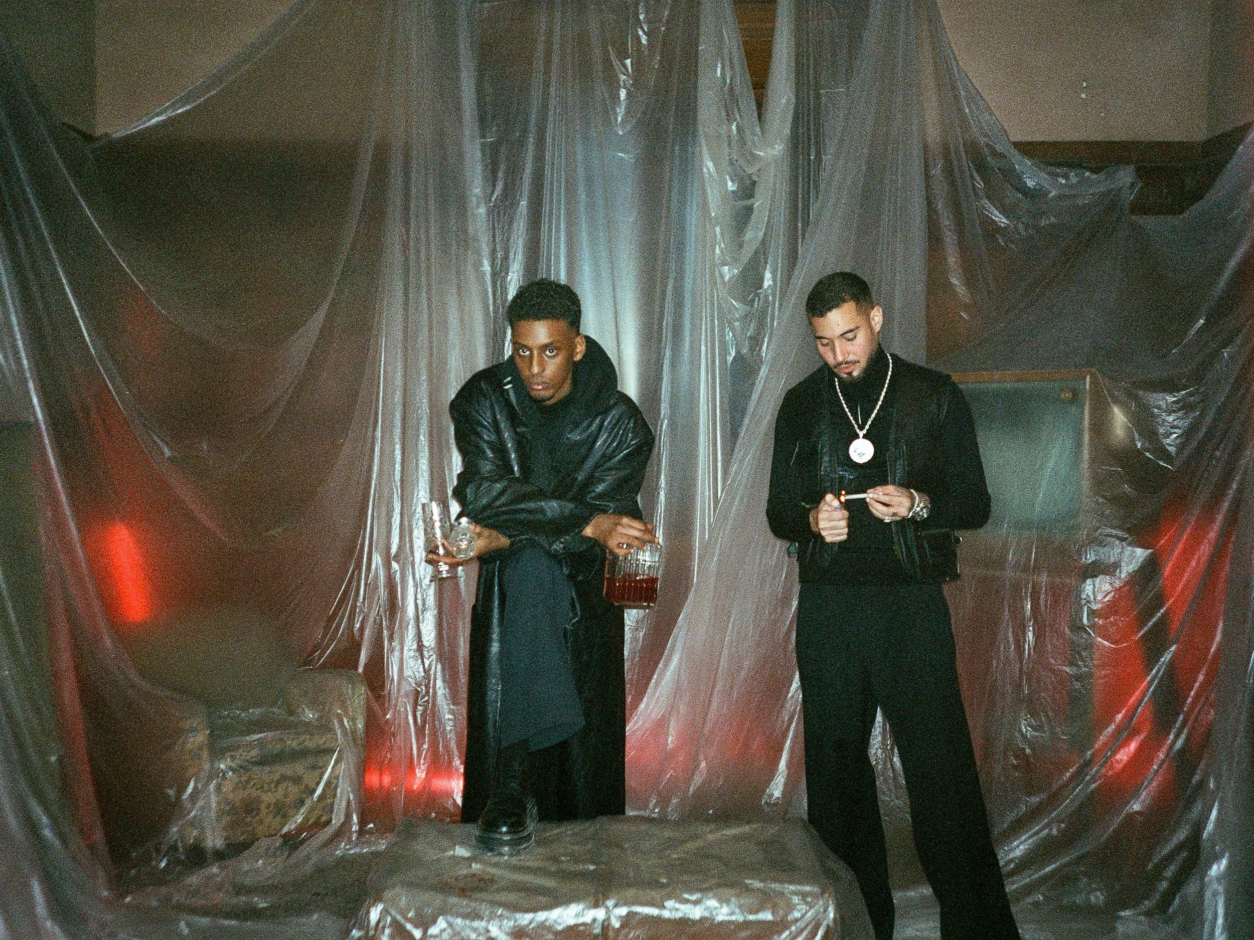 Yasin & Haval standing in front of a background covered in clear plastic sheets, one holding a glass and the other looking at a mobile device, both dressed in black athletic wear and jewelry.