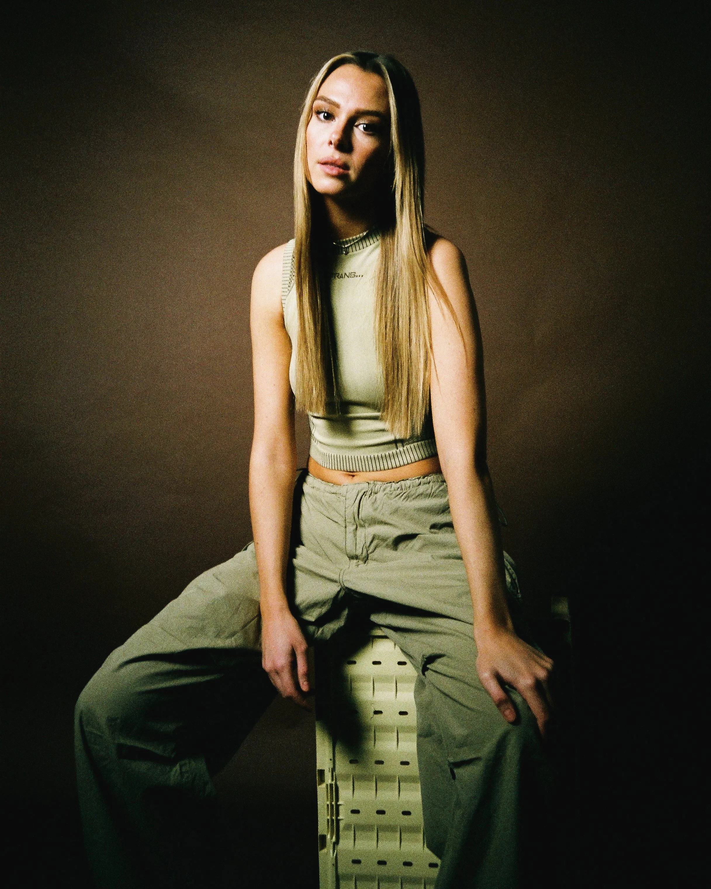 A Junie with long, straight blonde hair is sitting on a white plastic crate against a plain dark background, wearing a sleeveless beige top and loose beige pants, looking directly at the camera.