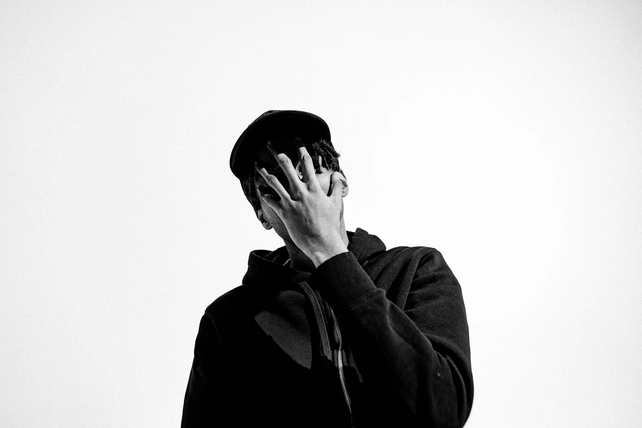 Black and white photo of a person wearing a cap and hoodie, covering their face with one hand, against a blank light background.