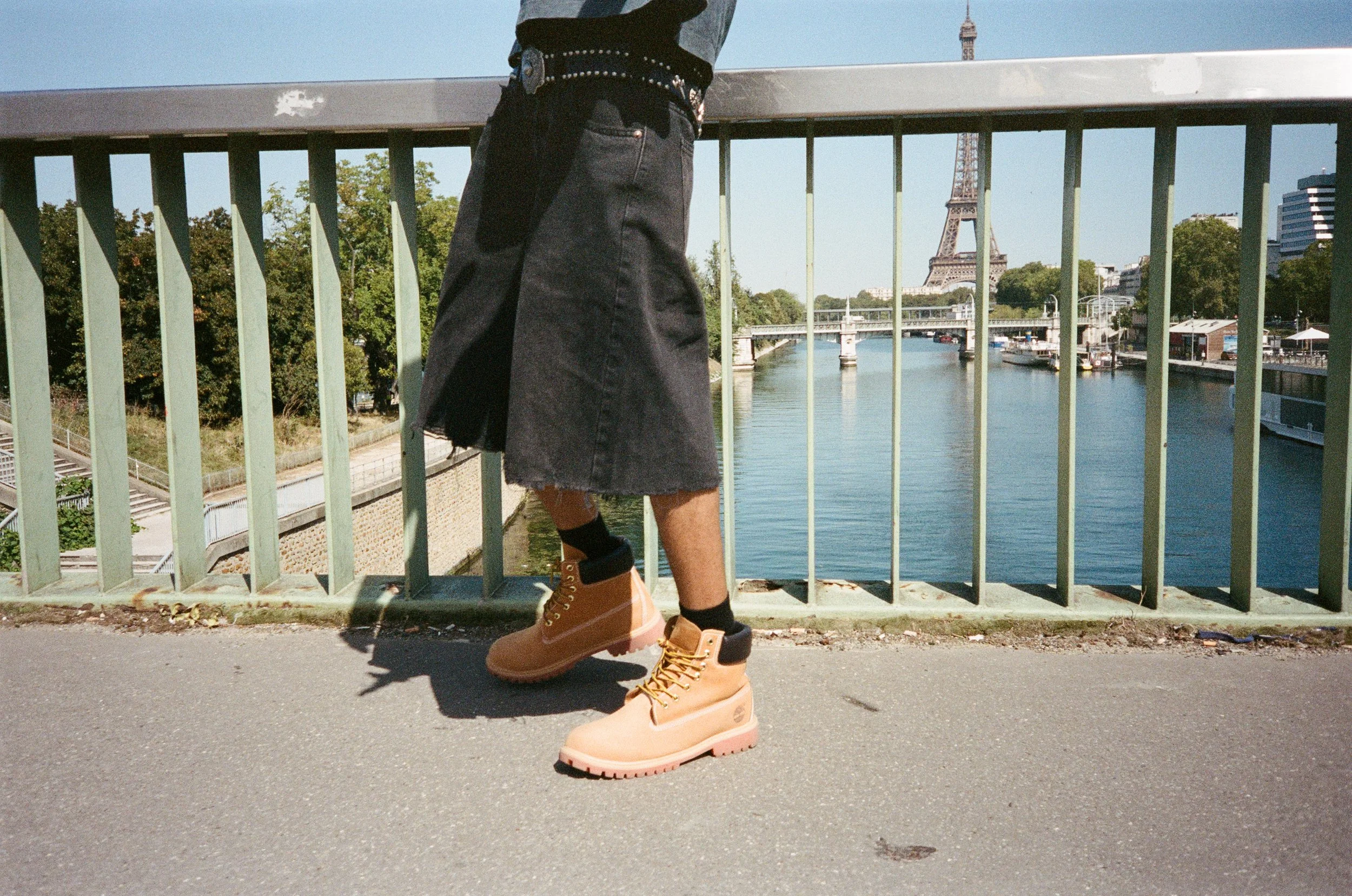Person standing on a bridge in front of the Eiffel Tower with a river below, wearing black shorts and beige boots.