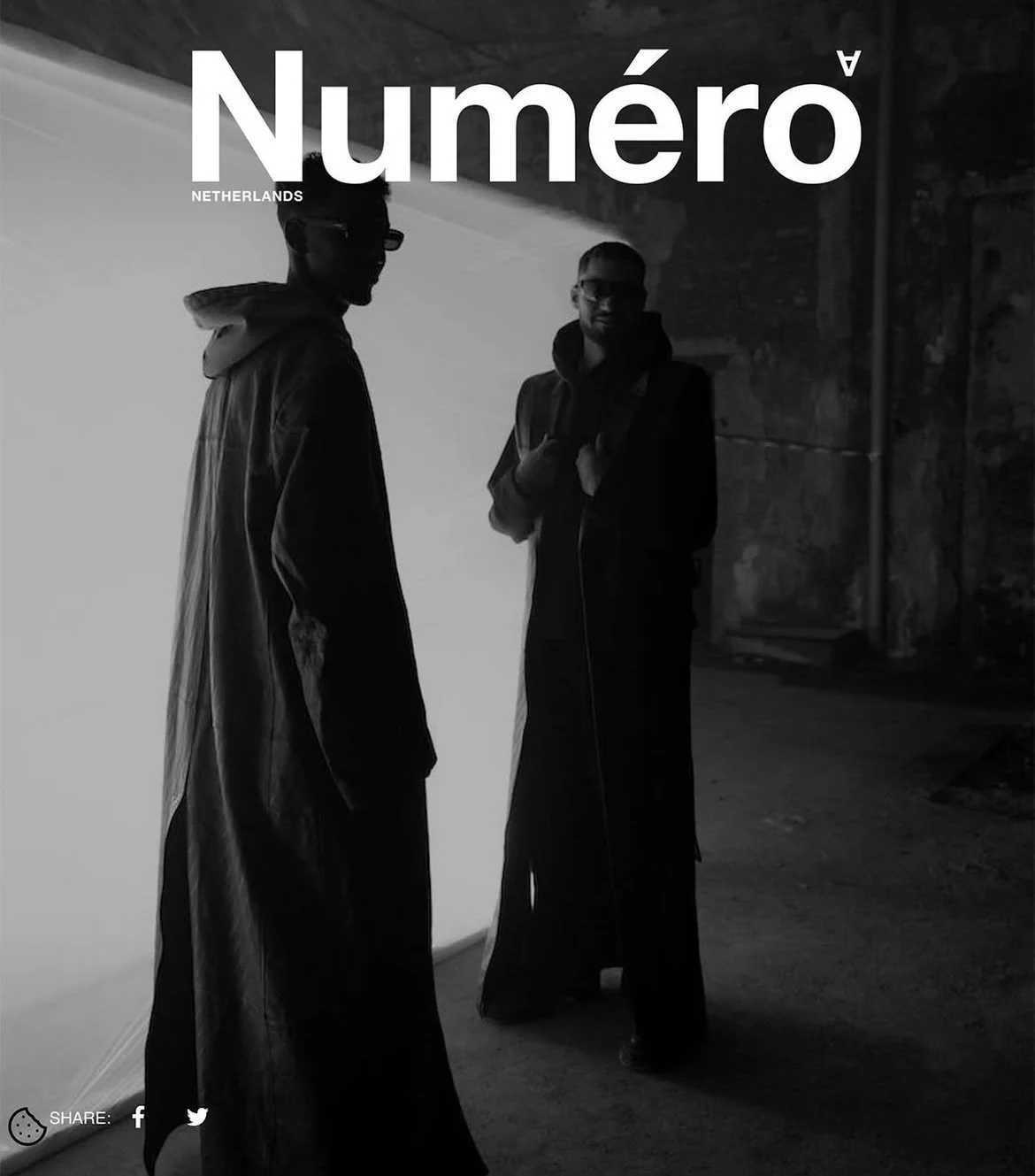 Black and white photo of two men standing and talking in an industrial or warehouse setting. One man is wearing glasses and a coat, the other is slightly in shadow. The text 'Numéro Netherlands' is overlaid at the top.