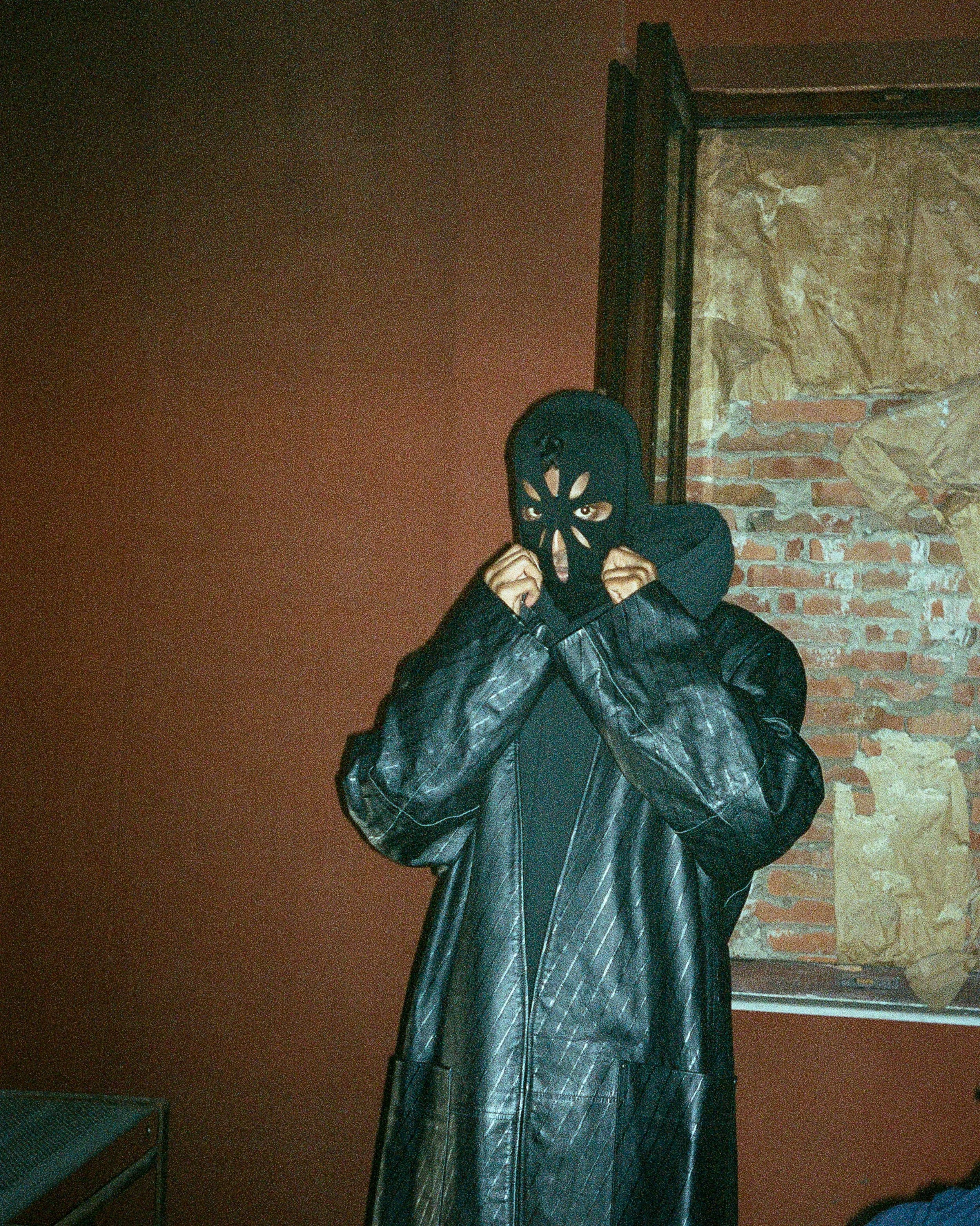 Person wearing a black ski mask and black leather jacket, standing indoors against a brick wall, adjusting the mask.
