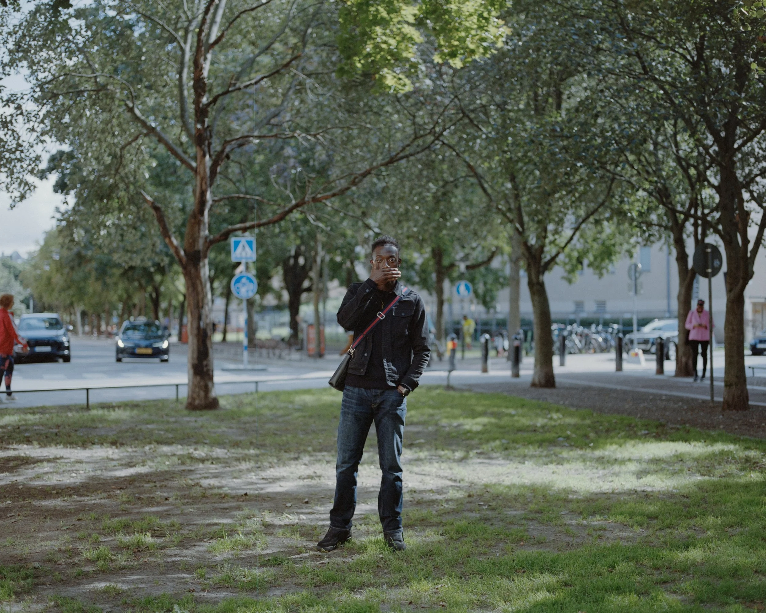 A young man standing on a grassy patch in a city park, surrounded by trees and traffic signs, with cars and pedestrians in the background.