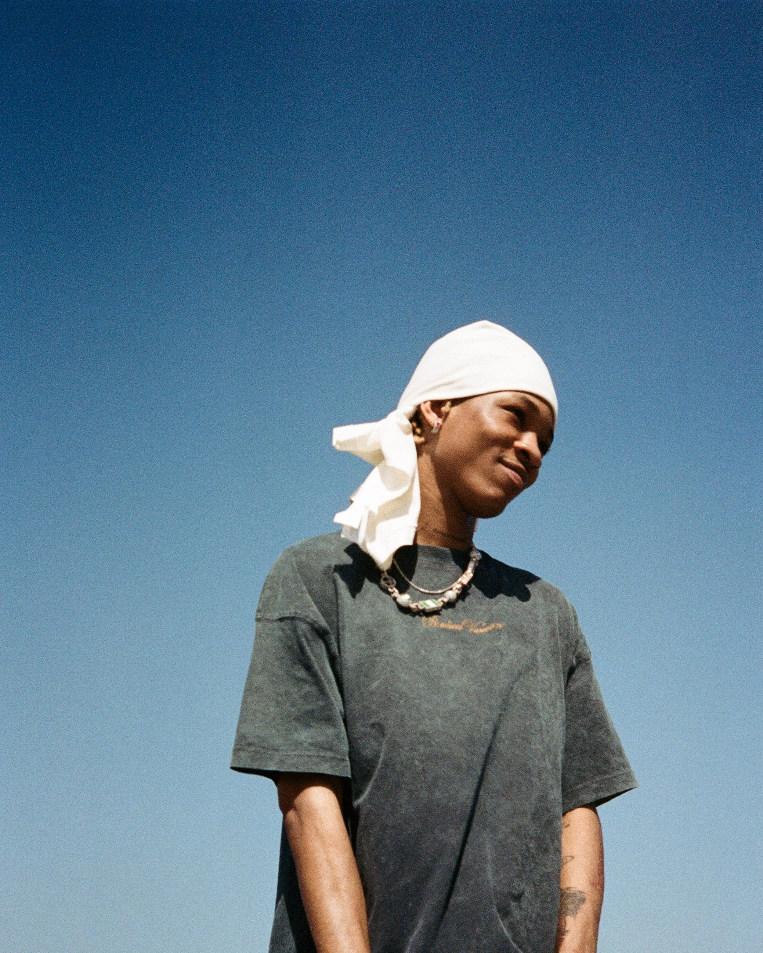 A person wearing a white headscarf, a dark T-shirt with a small logo, and layered necklaces, standing outdoors against a clear blue sky.