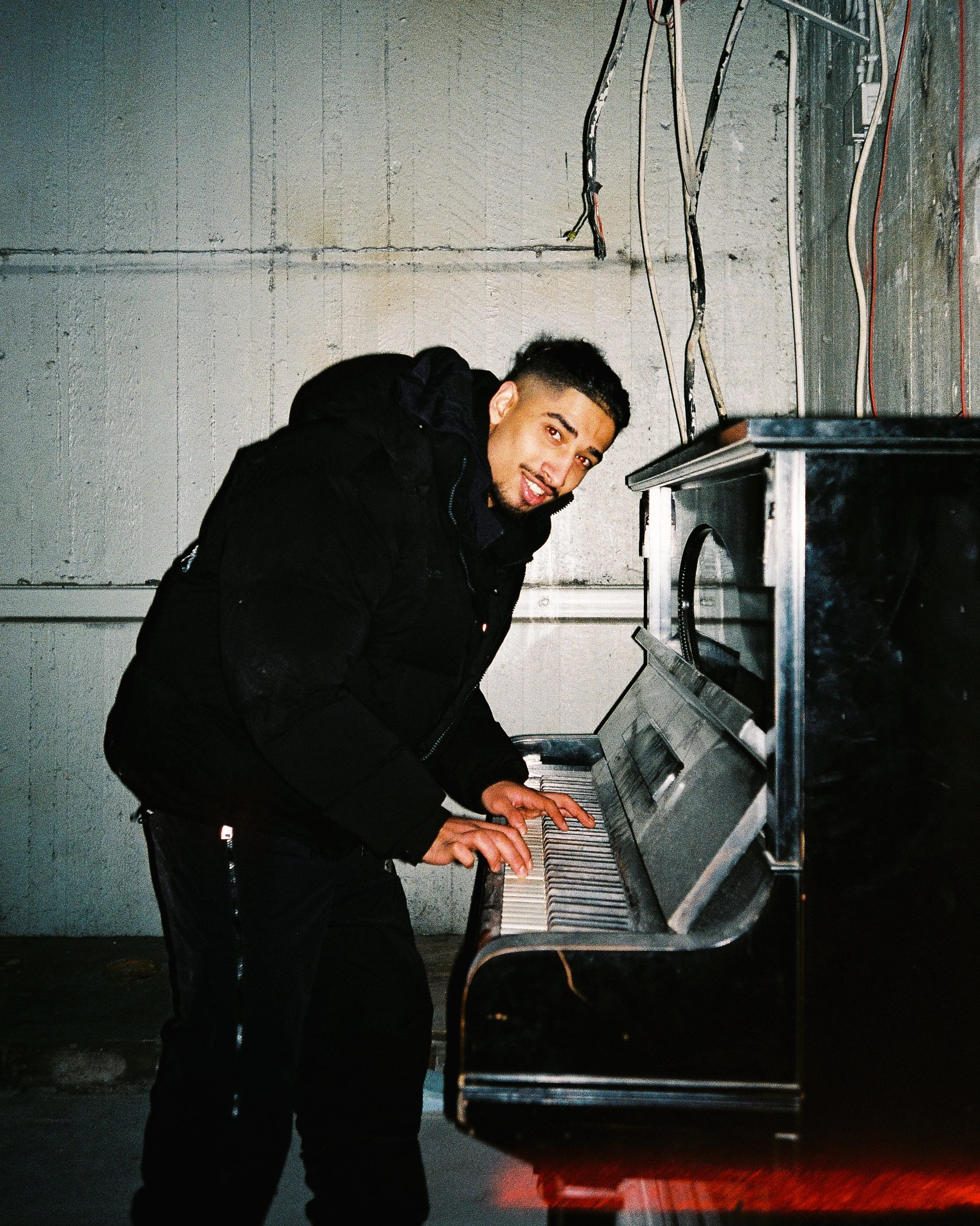 A young man in a black jacket playing an upright piano outdoors at night, with a brick wall and hanging electrical wires in the background.