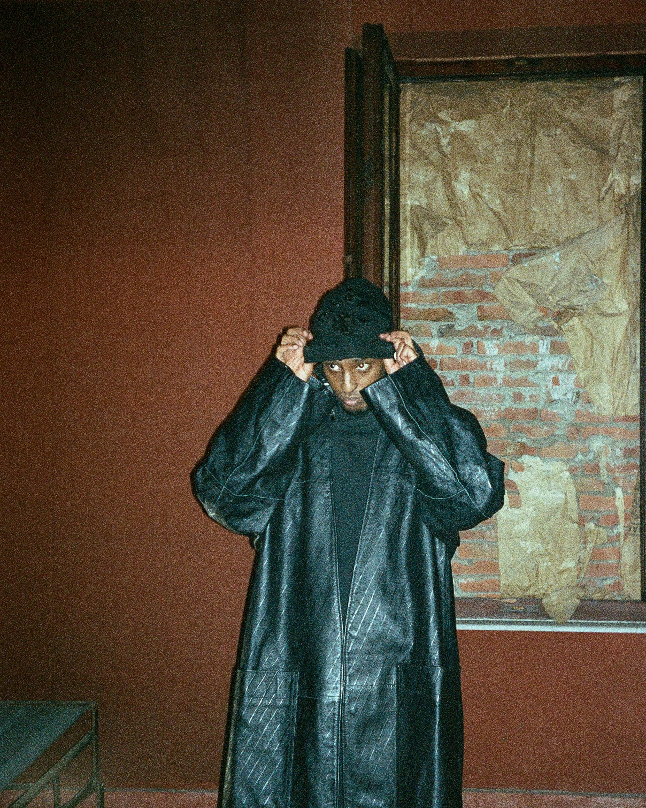 Yasin wearing a black leather coat and a black beanie, adjusting the beanie, standing indoors against a red wall with an abstract artwork.