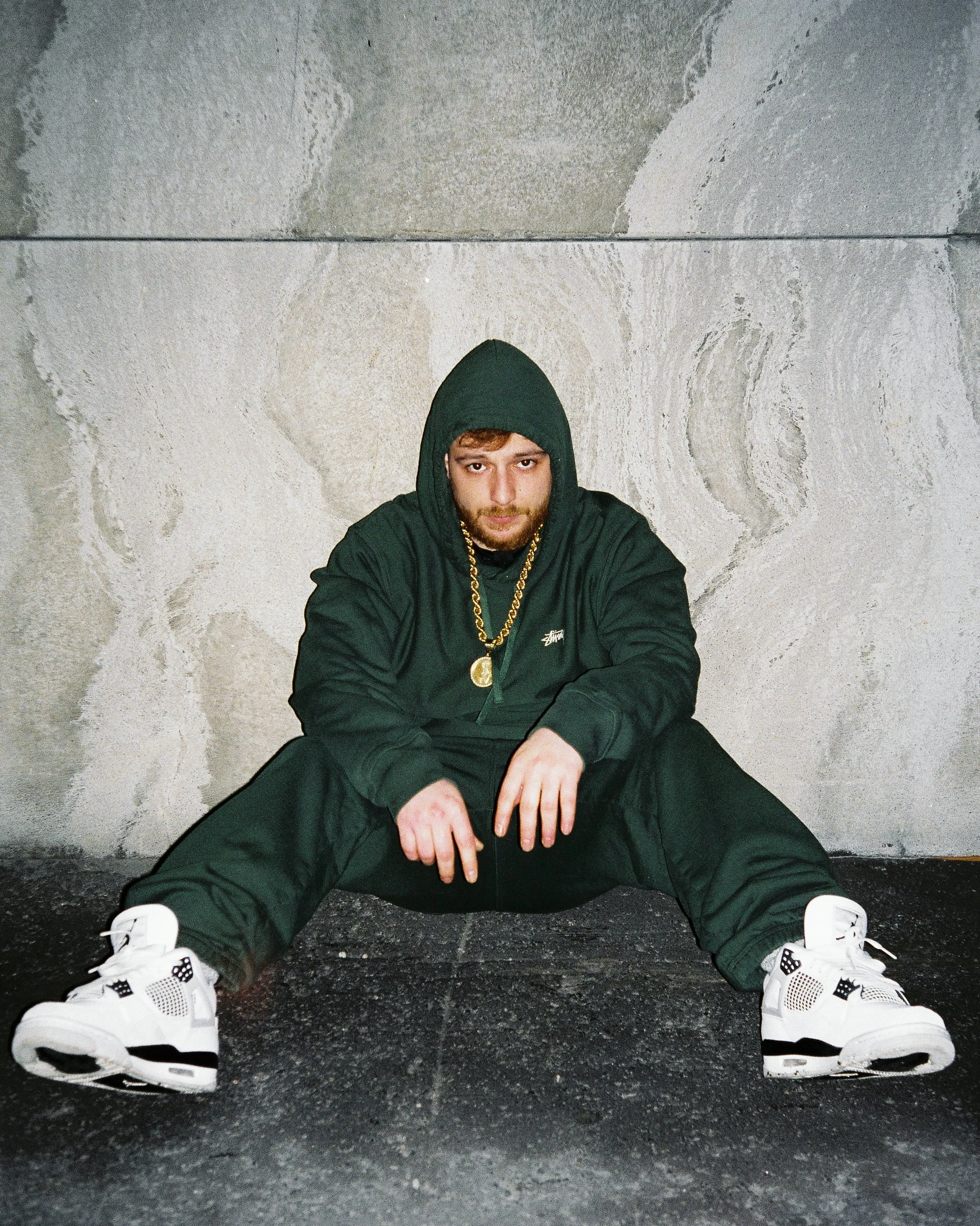 A man in a black hoodie, black pants, and white sneakers sitting on the ground against a concrete wall, wearing gold jewelry, with a serious expression.