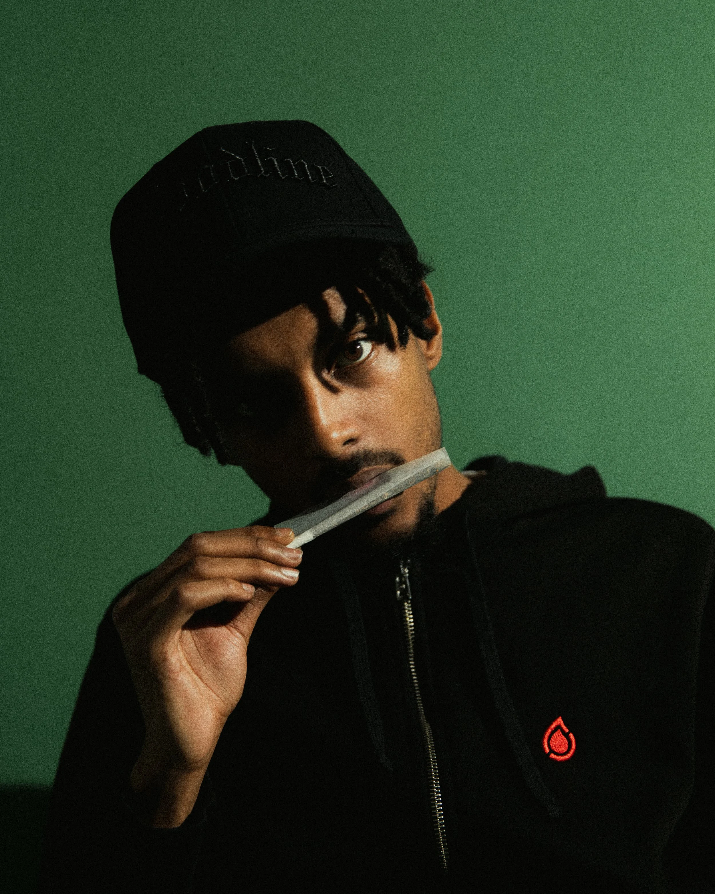 Young man with dreadlocks wearing a black baseball cap and hoodie, holding a dollar bill near his mouth against a green background.