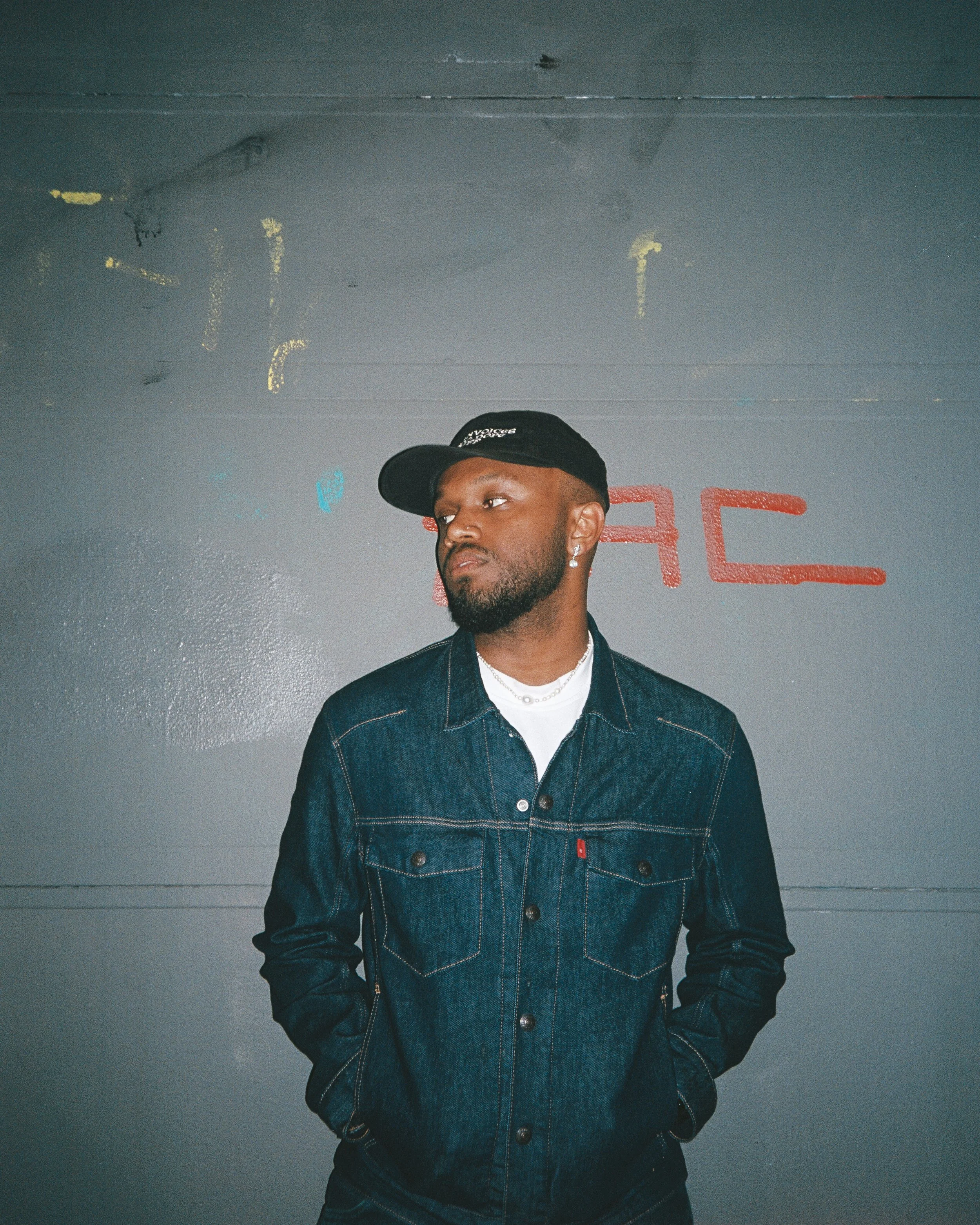 A man wearing a black cap, earrings, a denim jacket, and a white t-shirt standing in front of a gray wall with colorful graffiti.