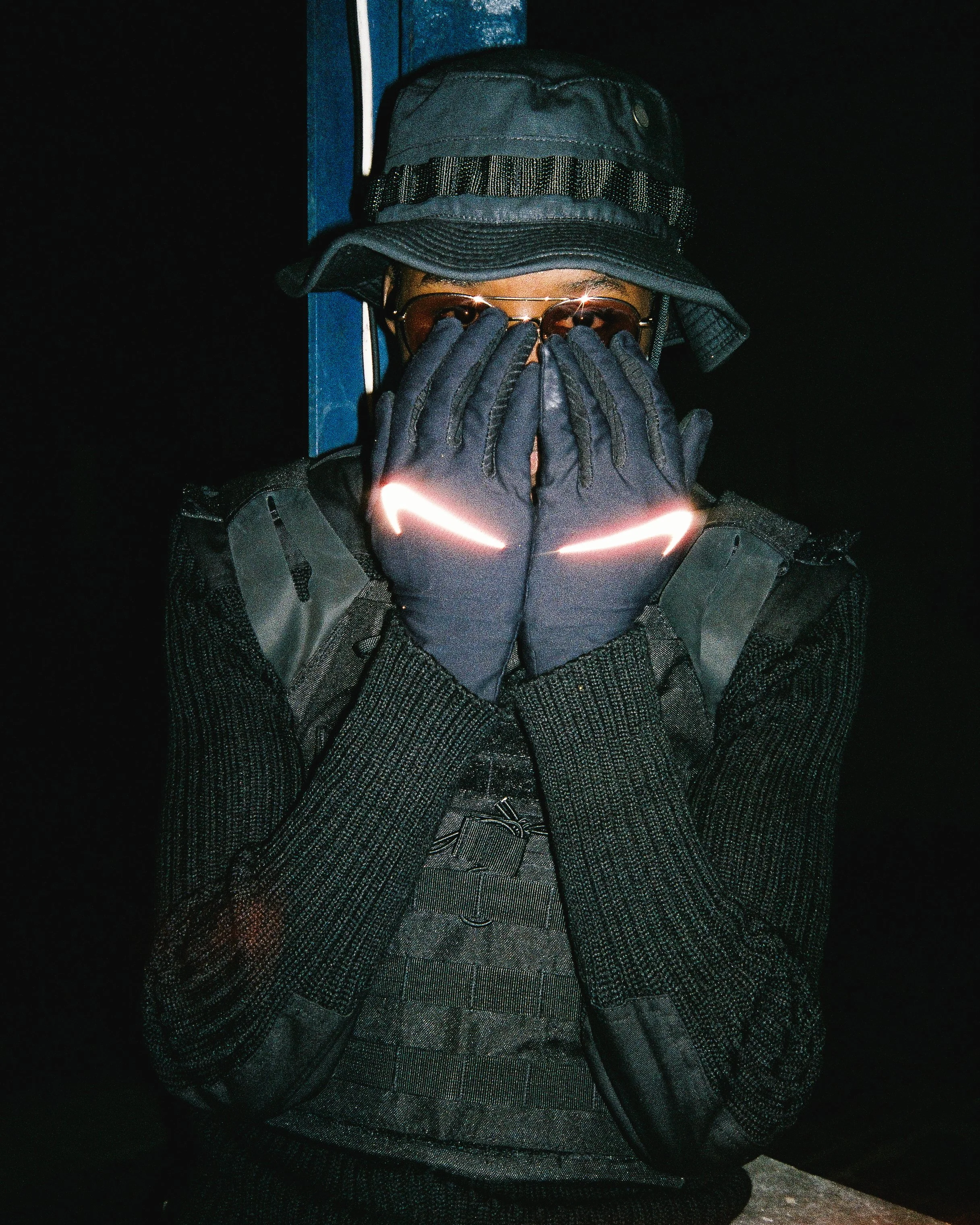 Youzie wearing a bucket hat, sunglasses, black gloves with reflective Nike logos, and a black jacket, covering their face with both hands.