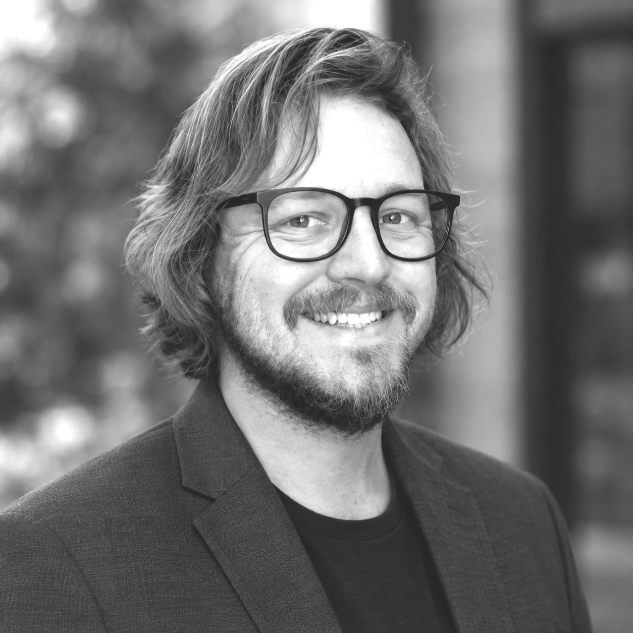 A smiling man with glasses, a beard, and a ponytail, sitting outdoors in front of bushes, wearing a polo shirt.